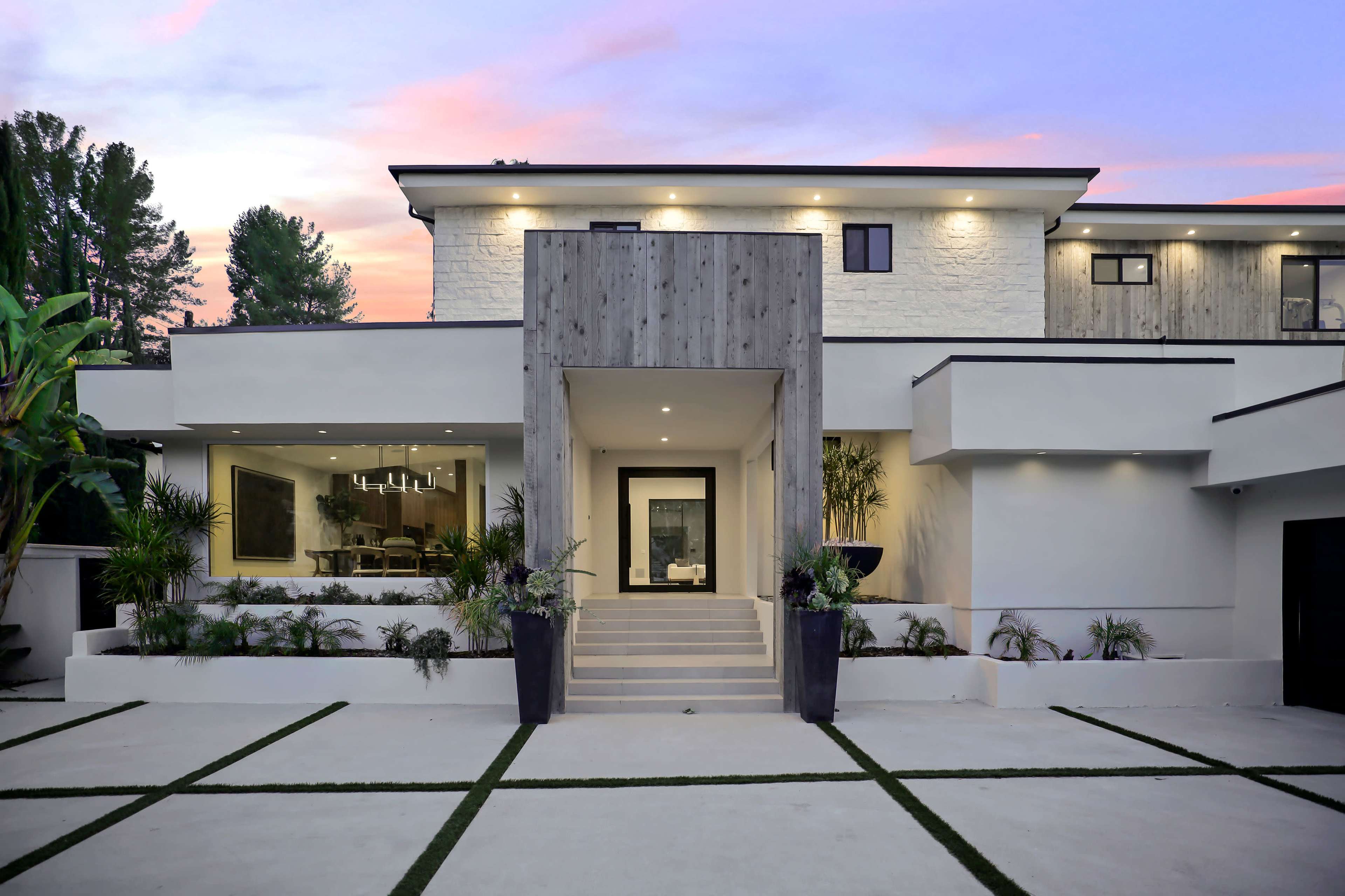 The image shows a modern two-story house with a large front entrance, surrounded by landscaping and a driveway leading up to it.
