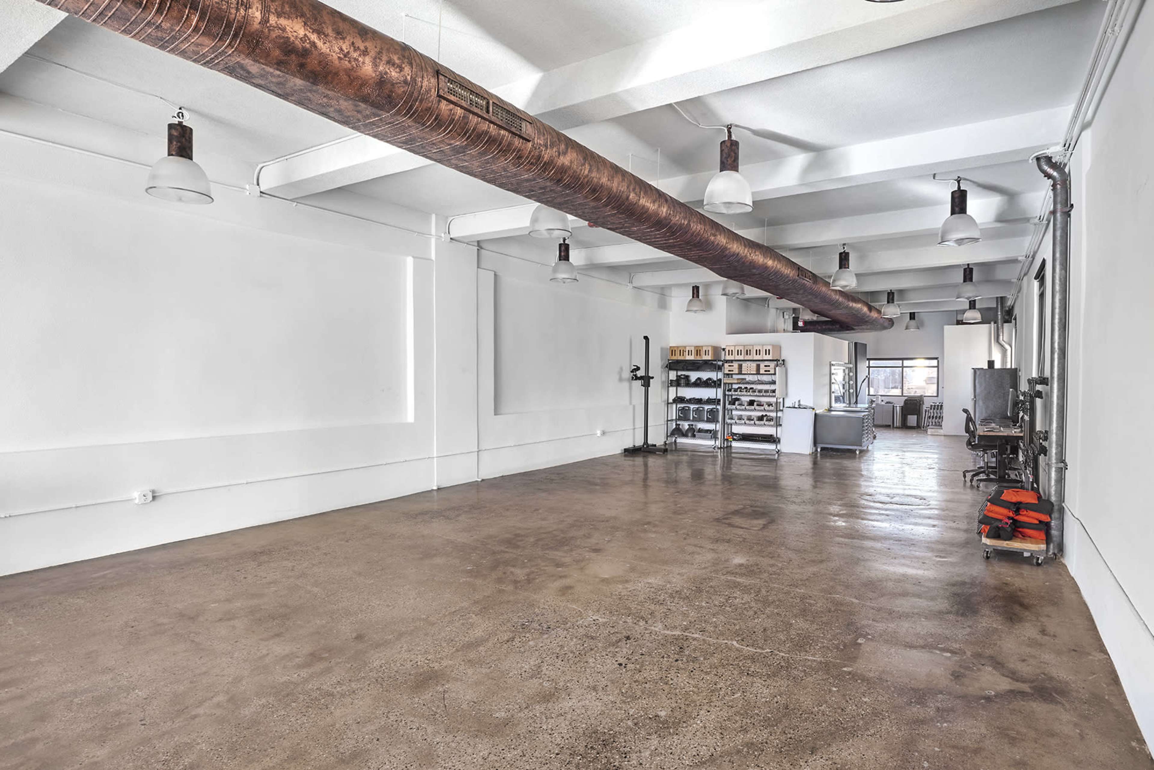 The image shows a spacious, empty room with polished concrete floors, exposed piping, and minimal furnishings.