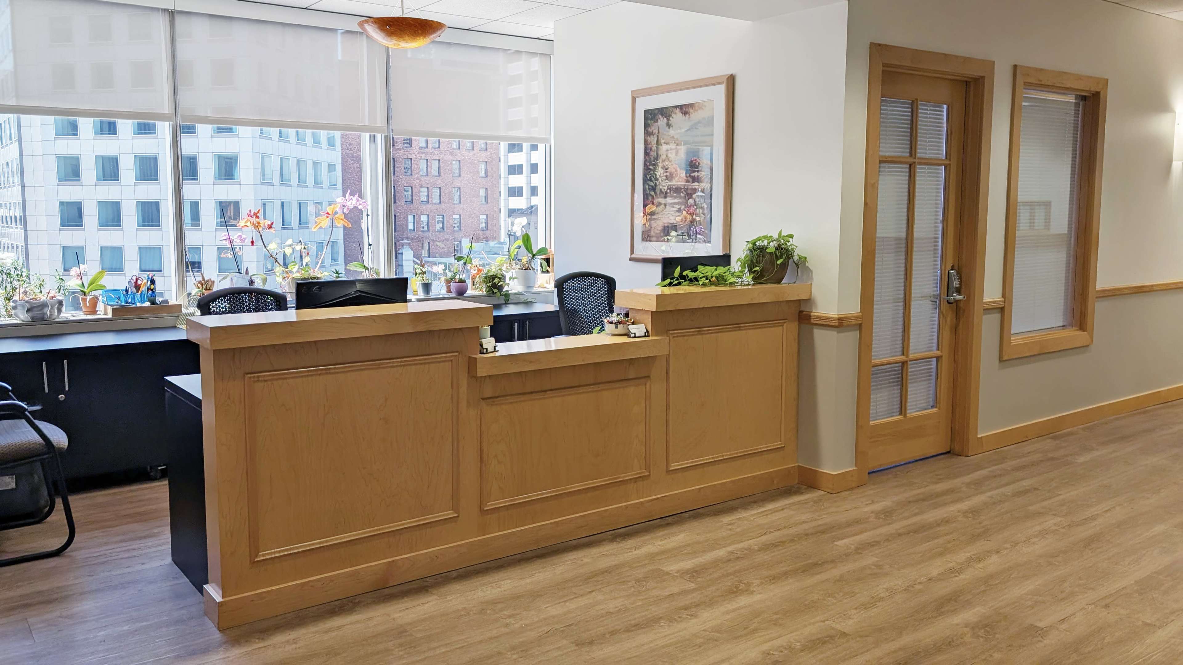 The image shows a reception area in an office with a wooden desk, plants on the counter, and large windows offering a view of a cityscape.