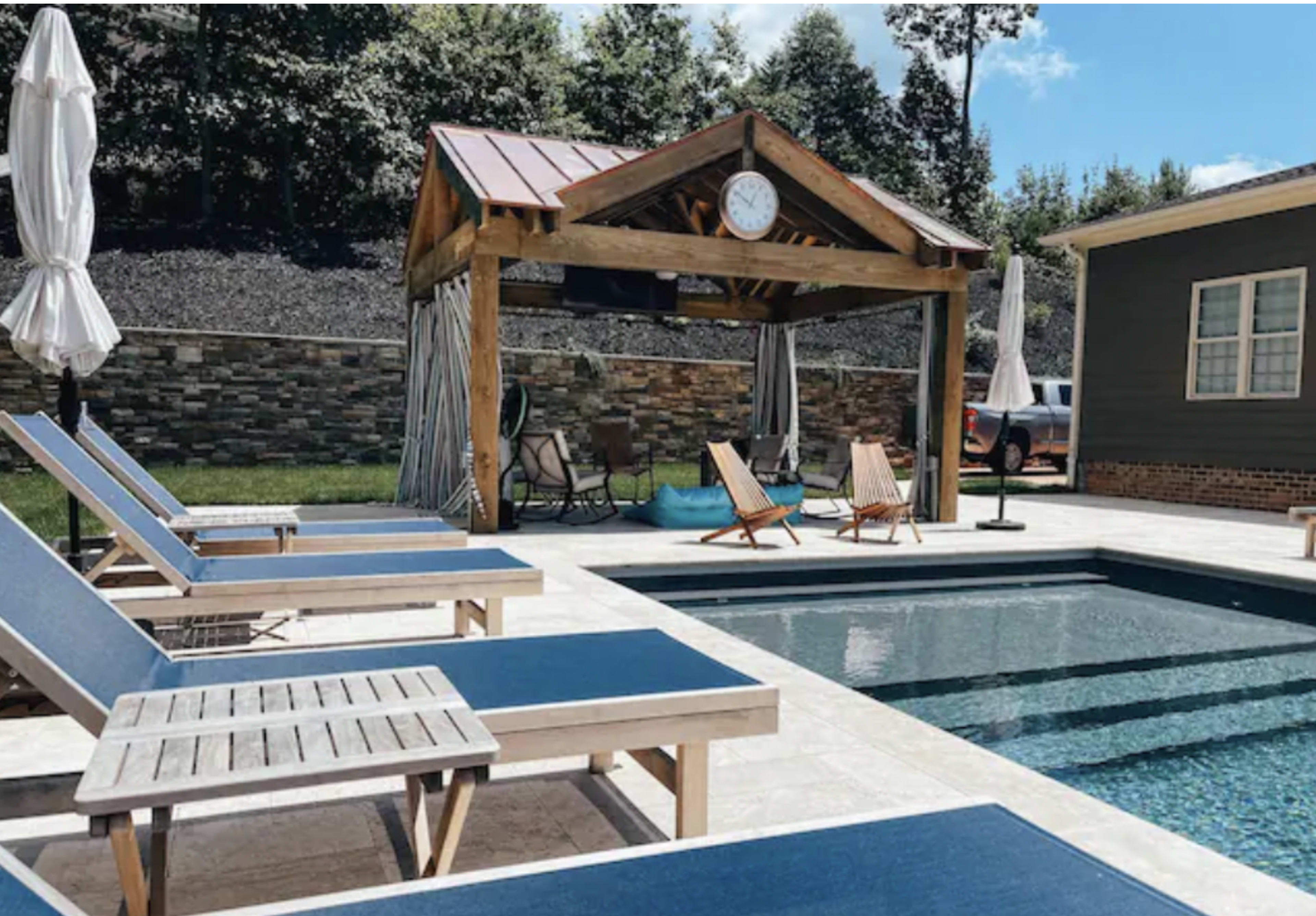 A swimming pool area features lounge chairs, a covered gazebo with a clock, and a stone wall backdrop.