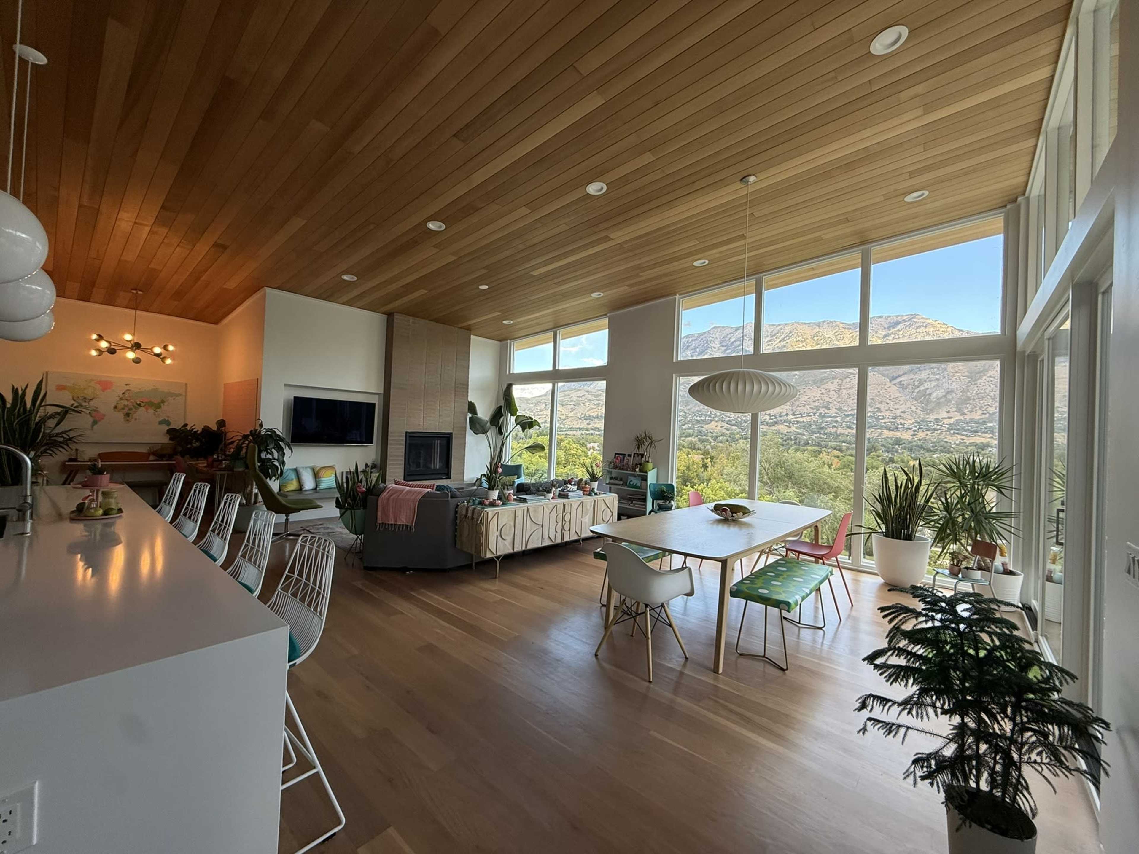The image shows a modern open-concept living space with large windows offering a view of mountains, featuring a dining area, a living area with a sofa, and various indoor plants.