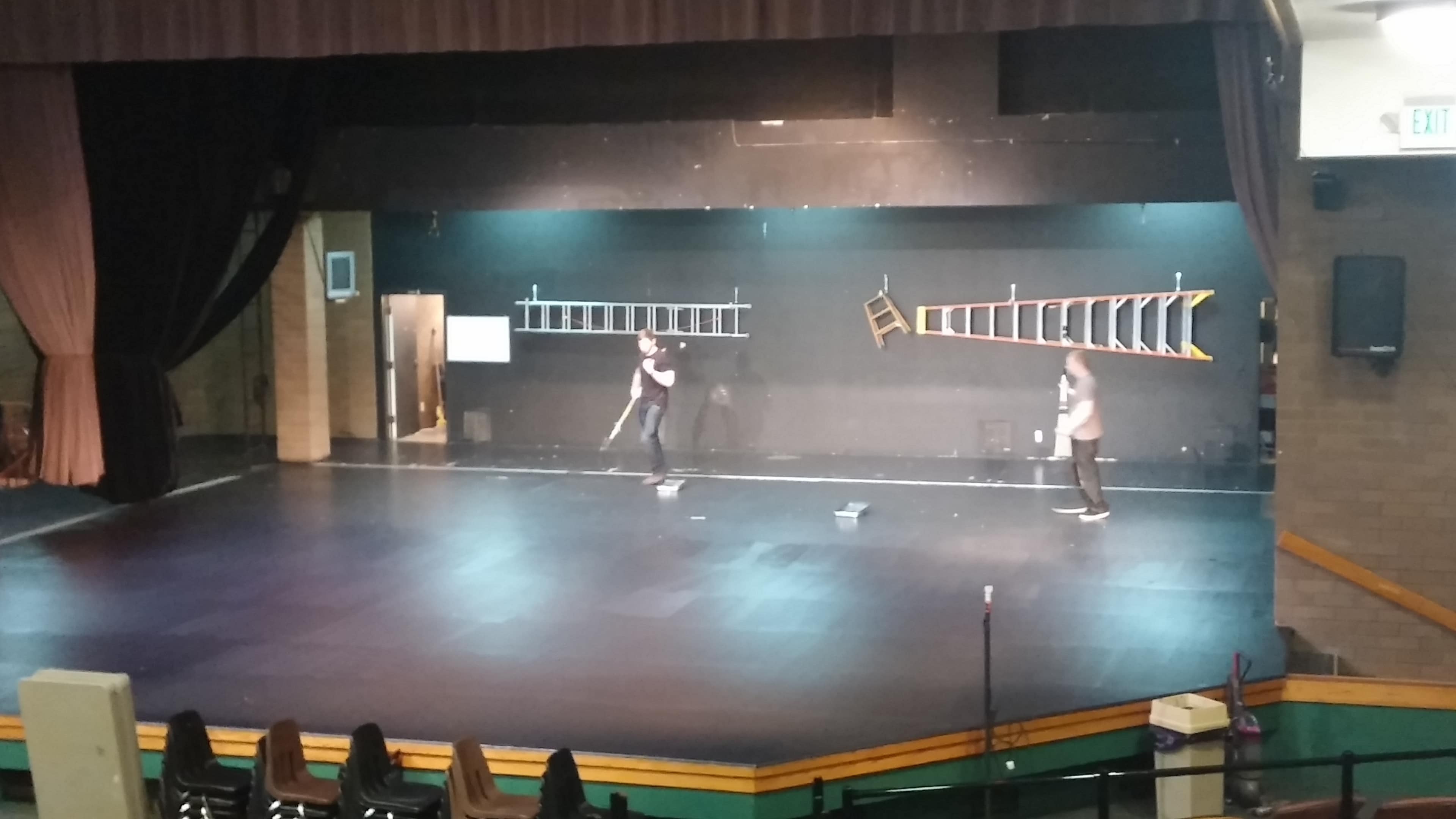 Two individuals are preparing a stage with ladders and a chair on a set lit by overhead lights.