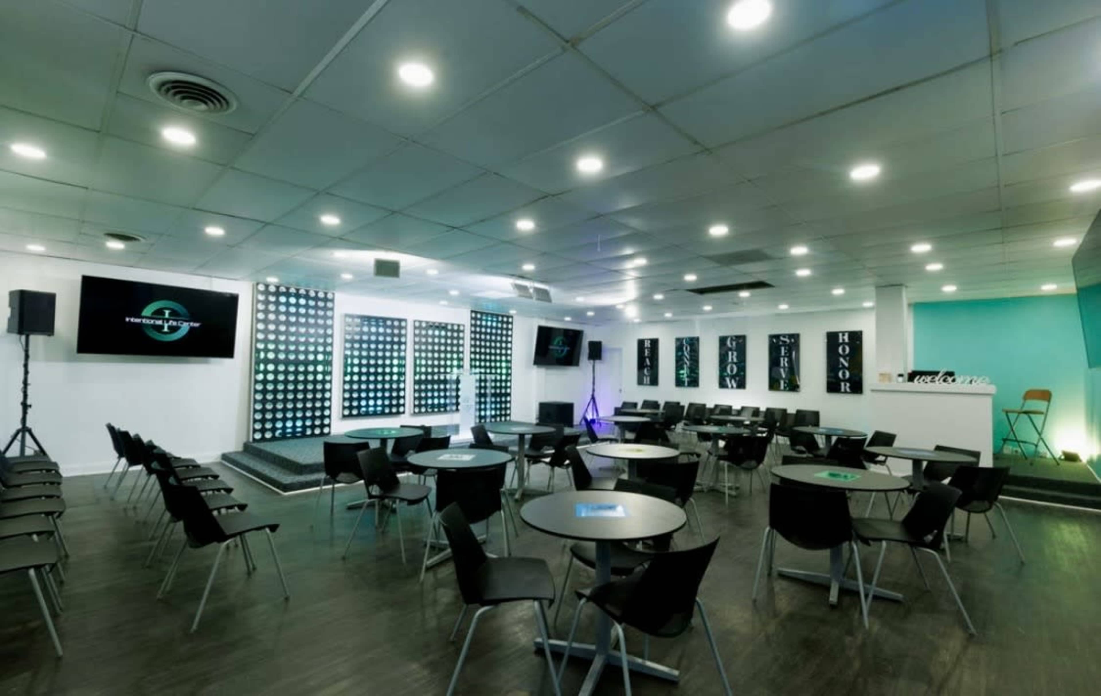The image shows a modern conference room set up with round tables and chairs facing a stage area, illuminated by bright lighting and decorated with a backdrop of lights.