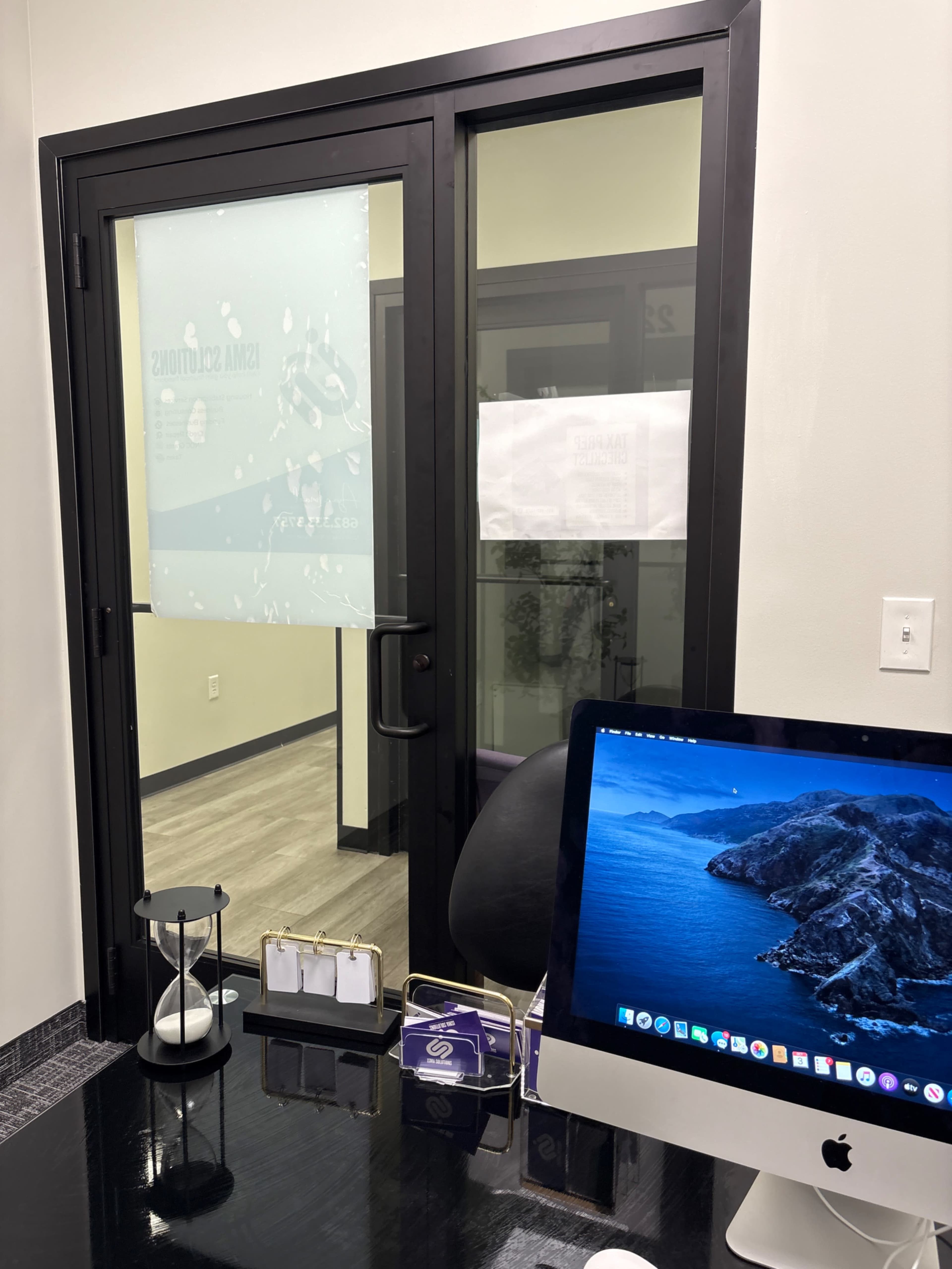 The image shows an office space with a glass door featuring partial branding, a black desk with a computer, and decorative items arranged on the surface.