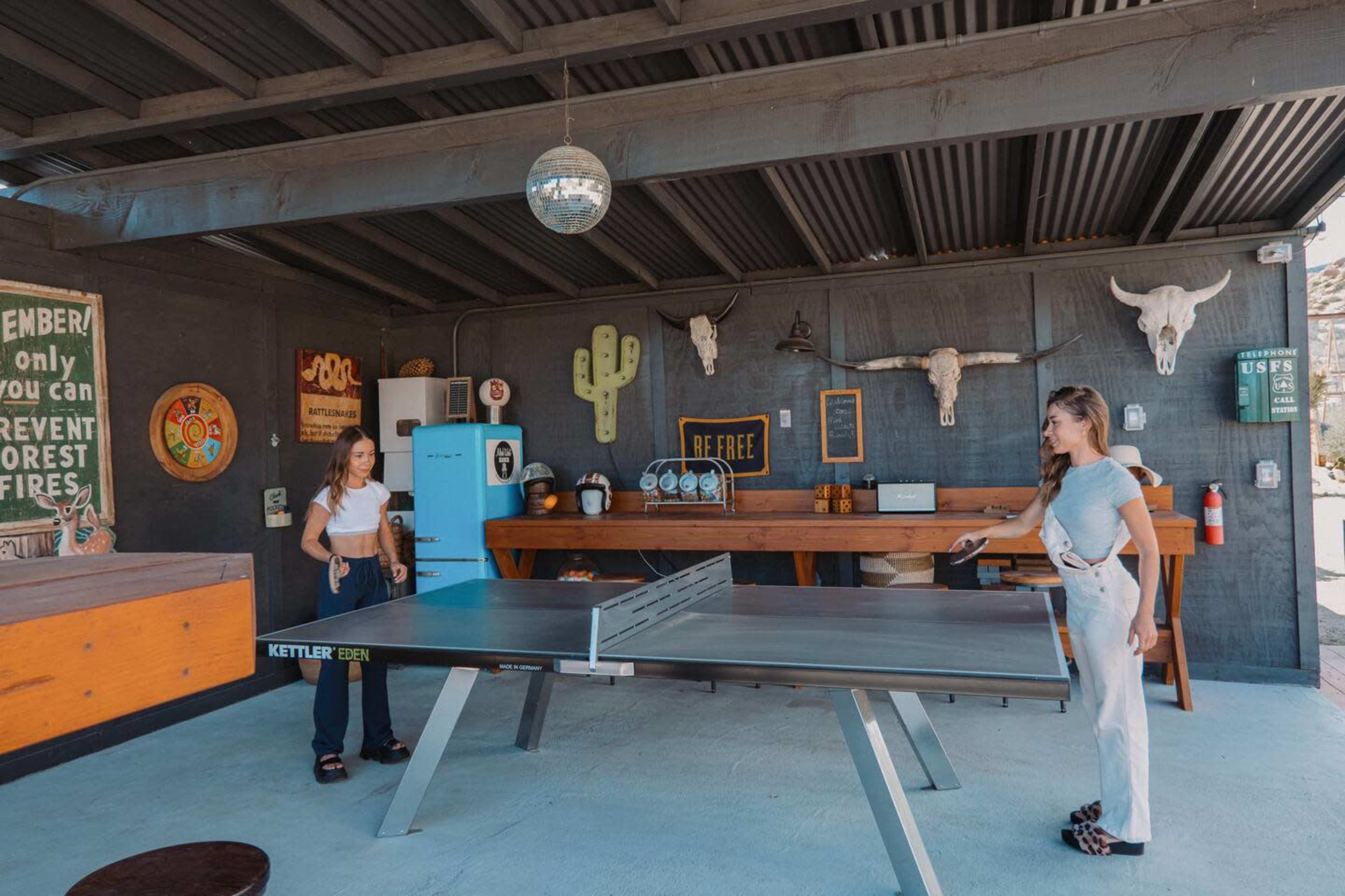 Two women are playing ping pong at a table set up in a rustic outdoor space decorated with western-themed artwork and decorations.