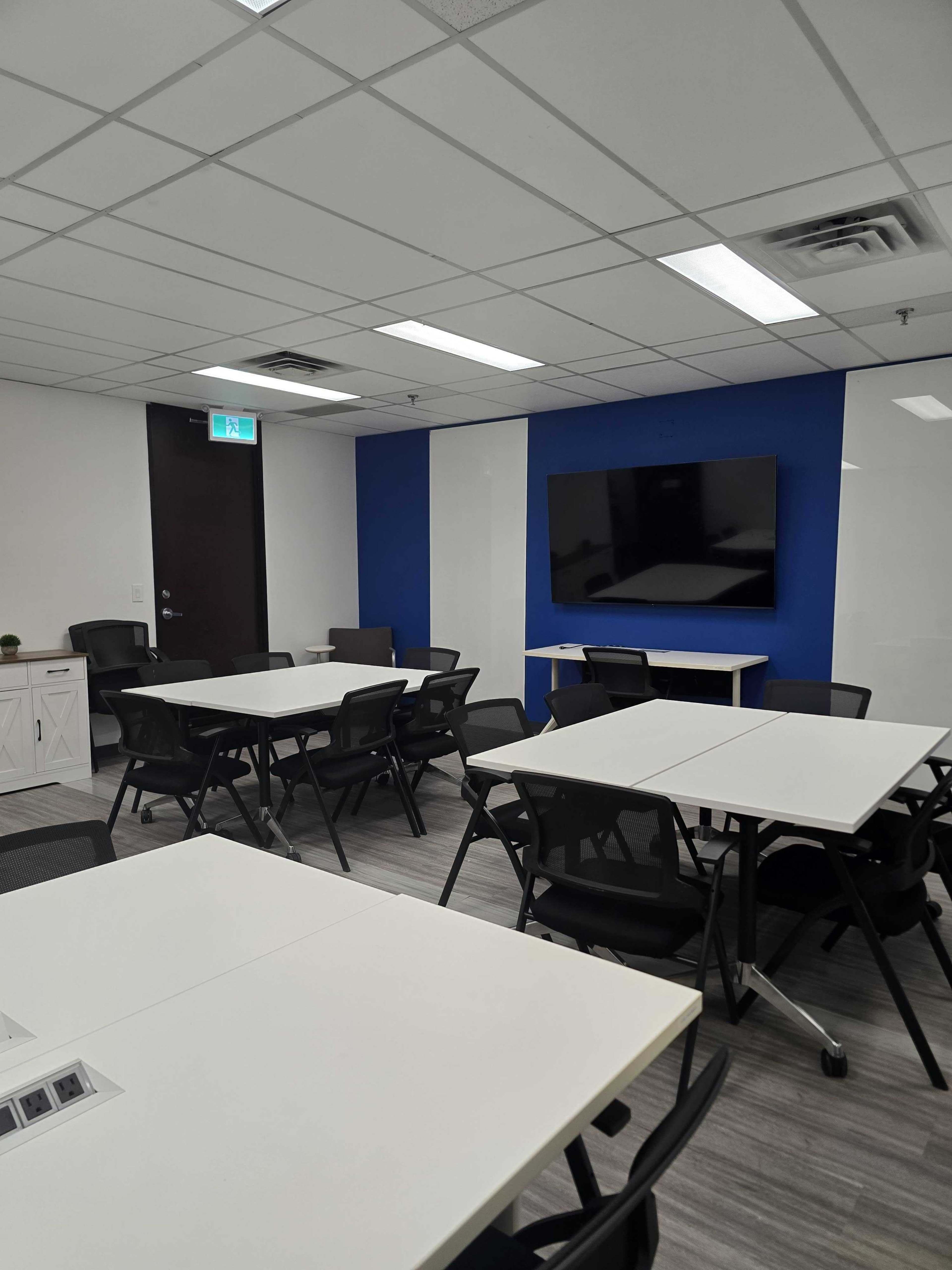 The image shows a modern conference room with multiple tables and black chairs arranged for meetings, featuring a large wall-mounted screen.