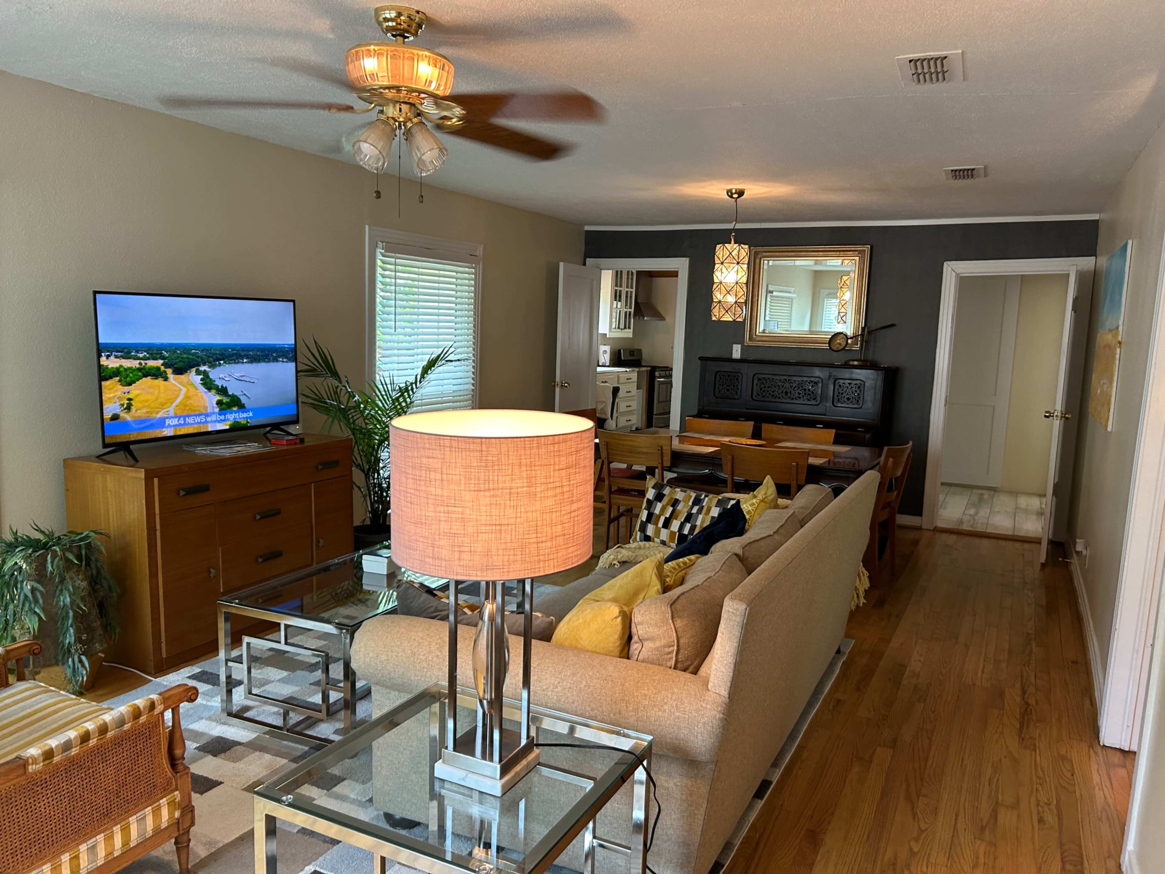 The image shows a living room with a beige sofa, a large television on a wooden cabinet, a coffee table, and a dining area in the background, illuminated by a ceiling fan and a lamp.