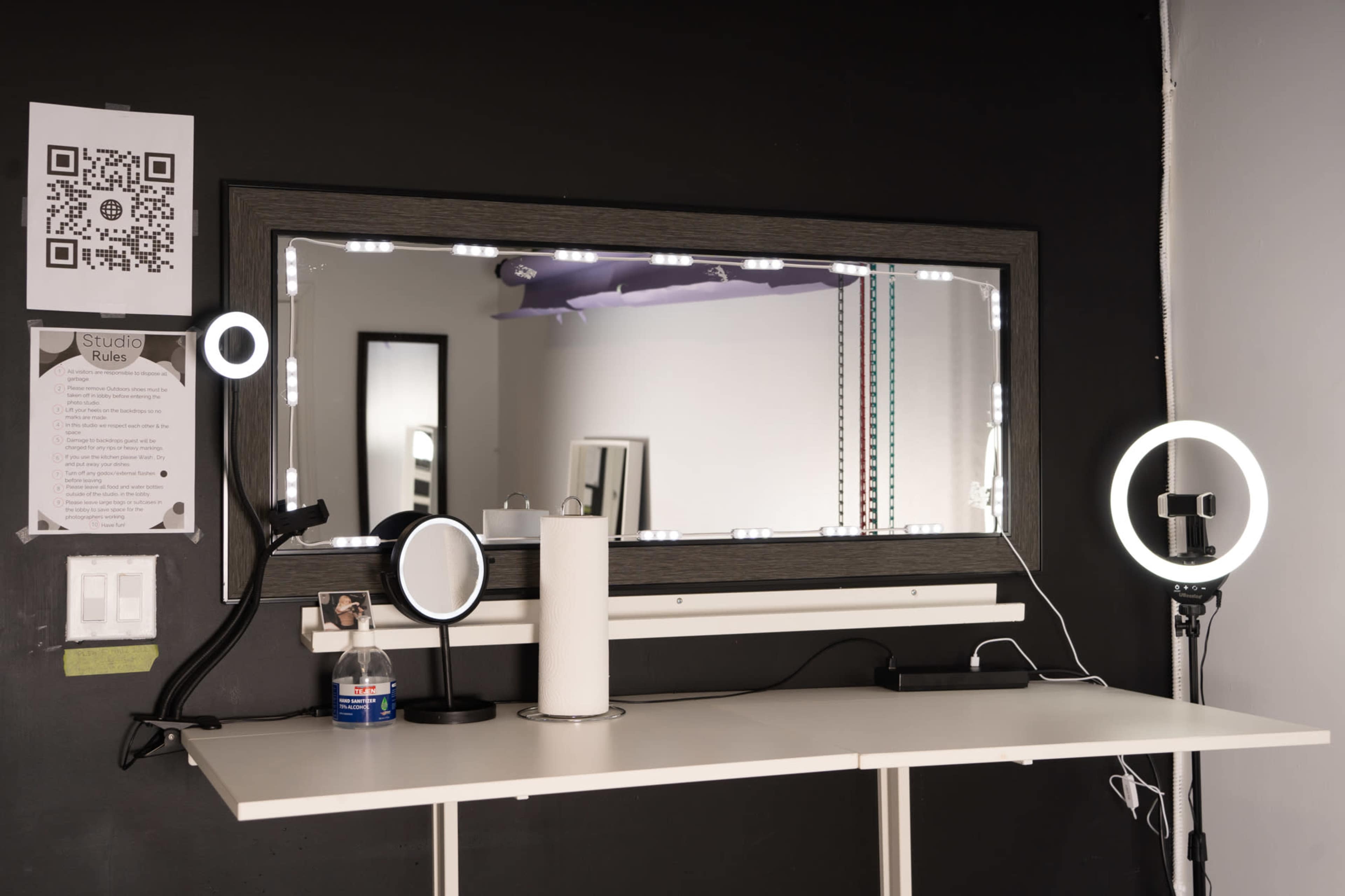 A vanity station with a large mirror, circular lights, and various devices on a white table against a dark wall.
