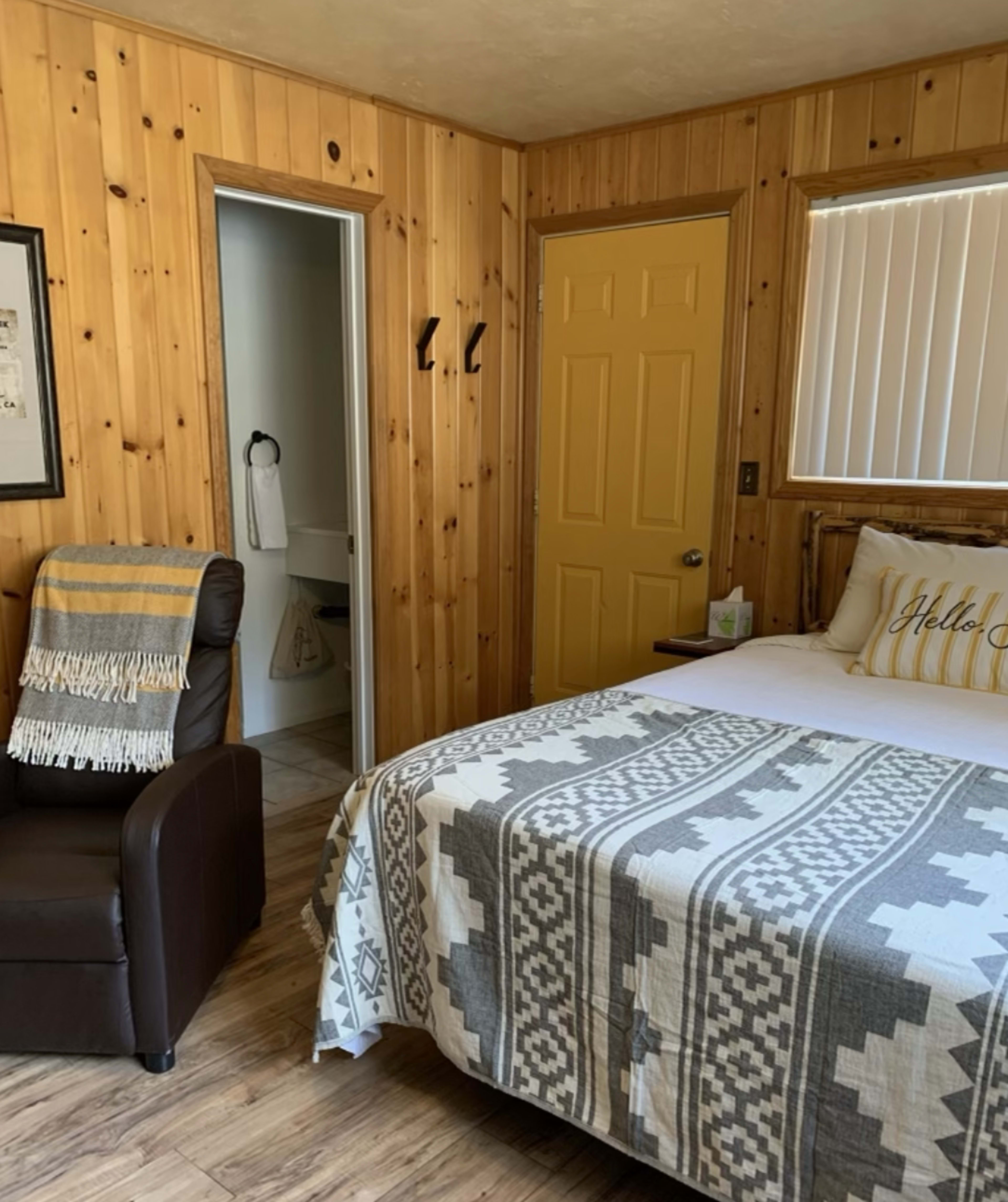 A small, cabin-style bedroom featuring a bed with a patterned blanket, a leather chair, wooden walls, and a yellow door leading to a bathroom.
