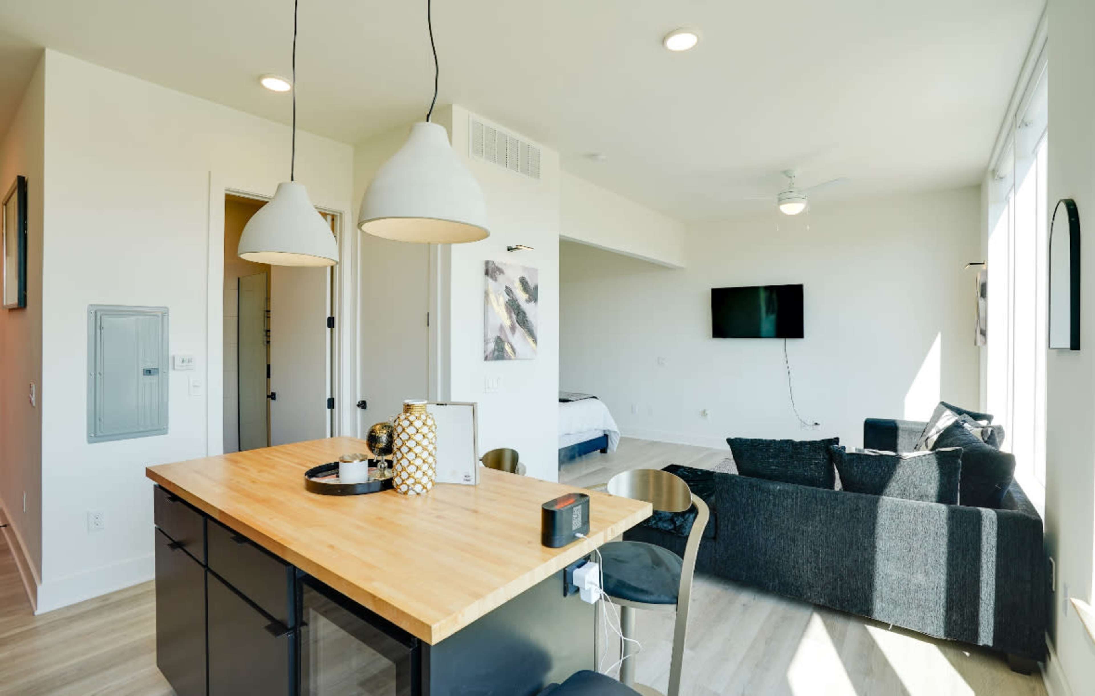 A modern living space featuring a kitchen island with pendant lights, a seating area with a black couch, and a wall-mounted television.