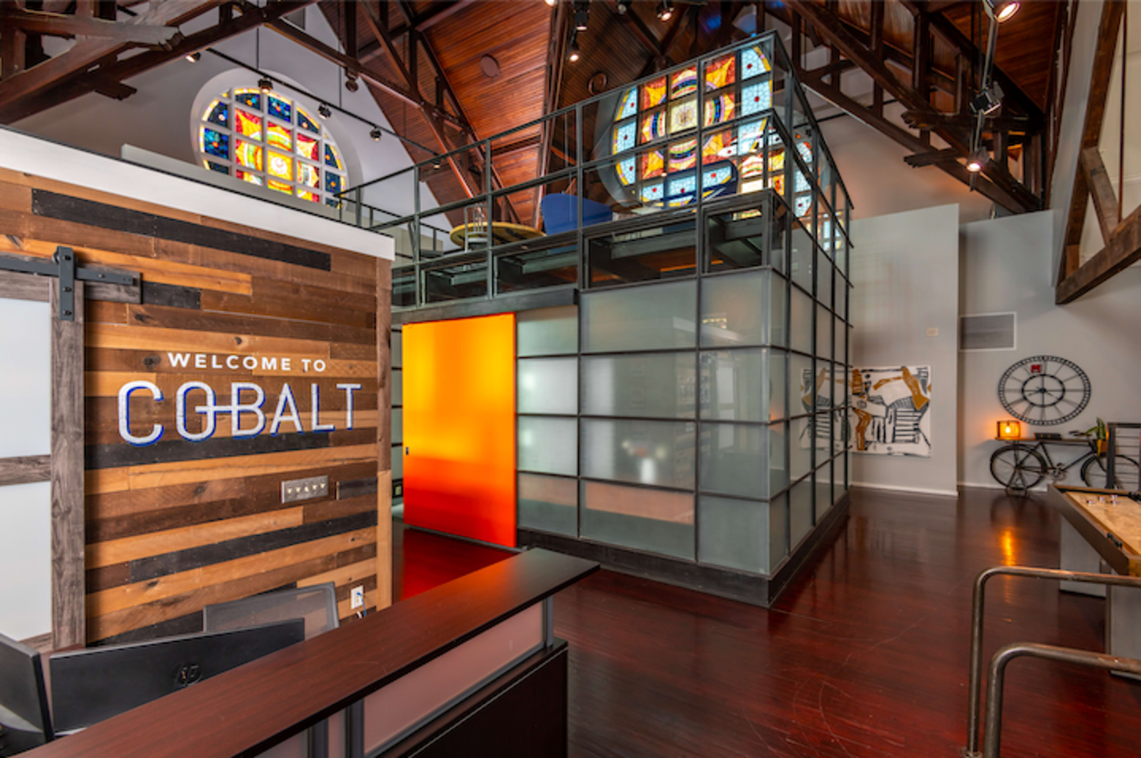 A modern office interior with a wooden reception area and glass-enclosed workspace, featuring colorful stained glass windows overhead.