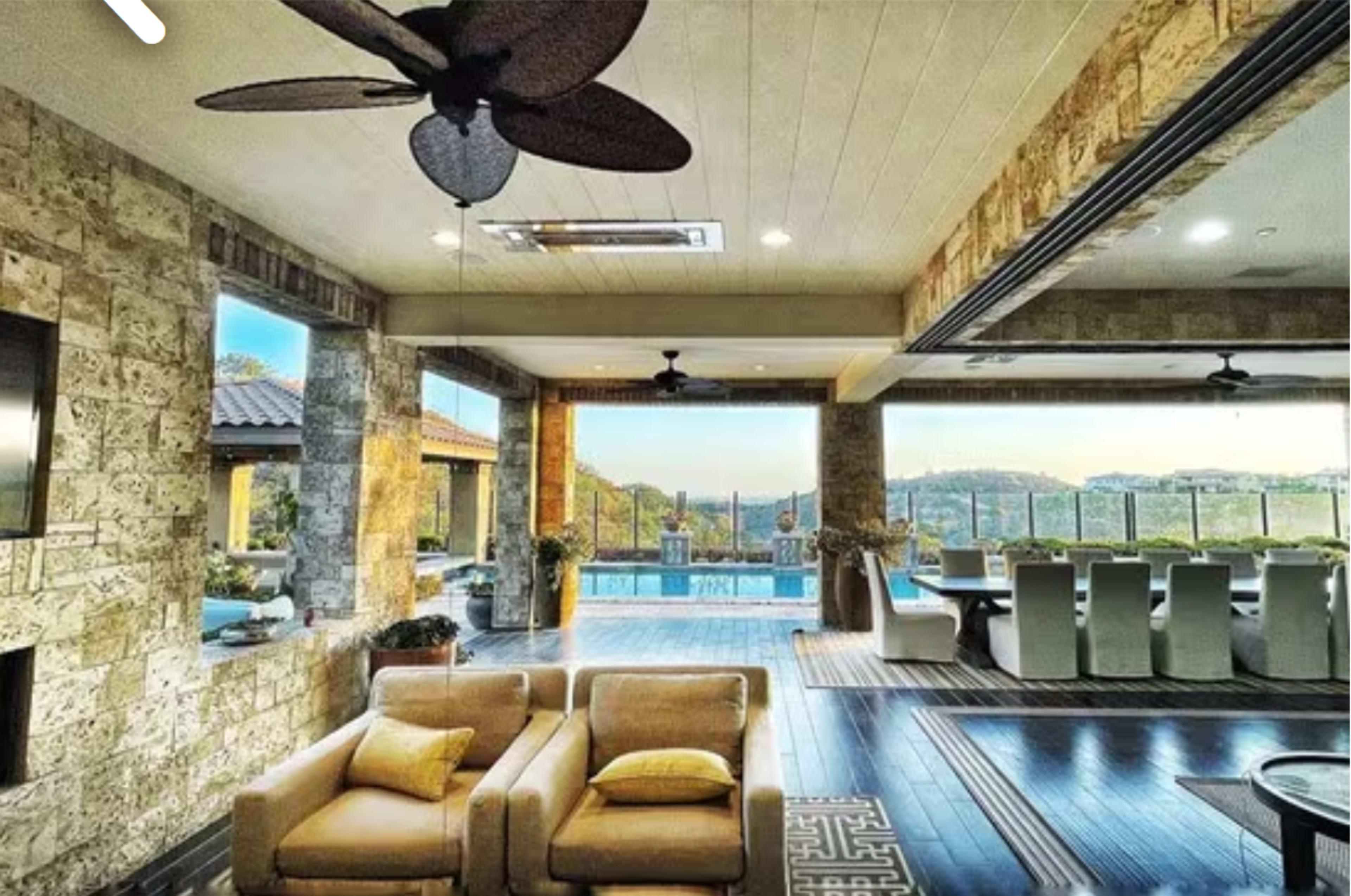 The image shows a spacious living area with stone walls, large windows overlooking a pool, and modern furniture.