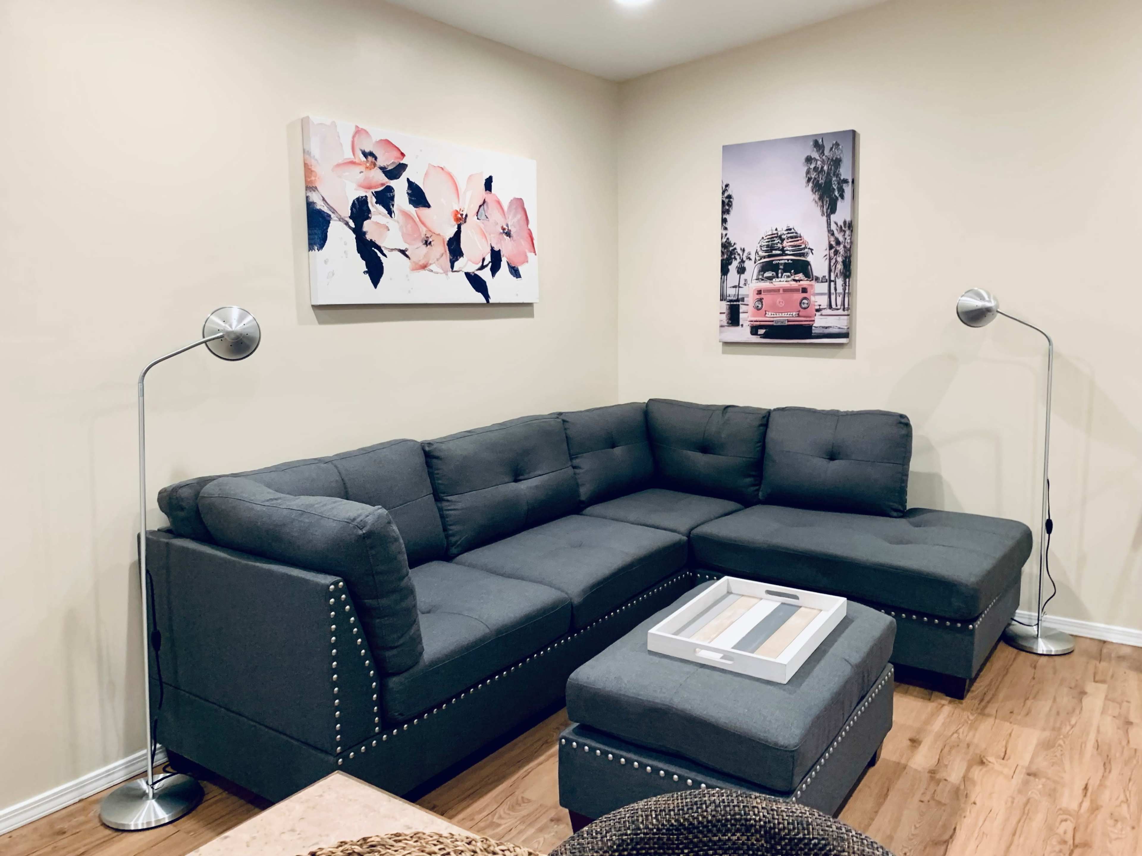 The image shows a modern living room featuring a gray sectional sofa, a coffee table, two floor lamps, and wall art depicting flowers and a vintage van.