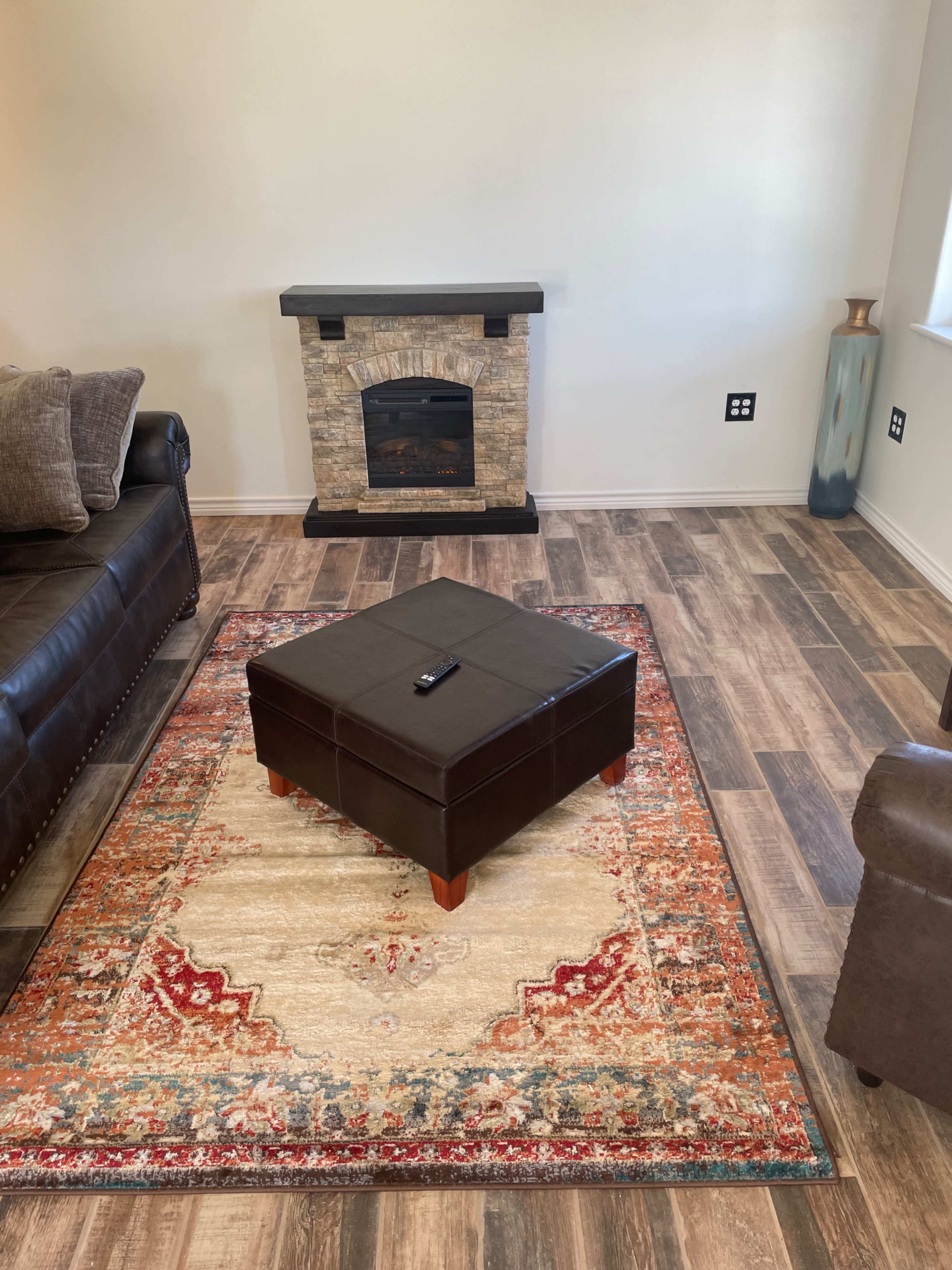 A living room with a stone fireplace, a brown leather sofa, an upholstered ottoman in the center, and a patterned rug on wooden flooring.