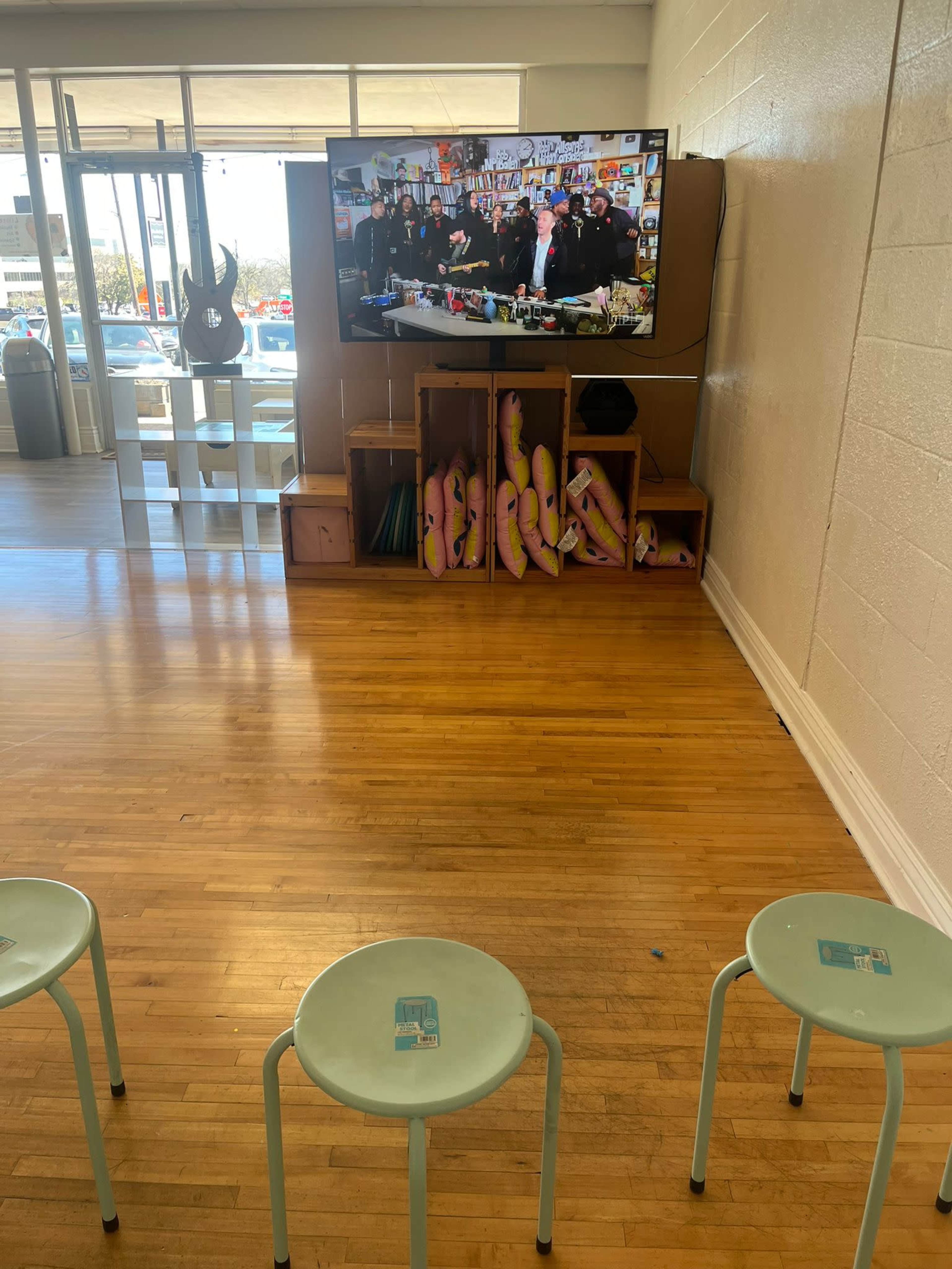 A room with wooden flooring features three metal stools facing a wall-mounted television displaying a group of people, alongside a shelf holding various items and some pink inflatable decorations.