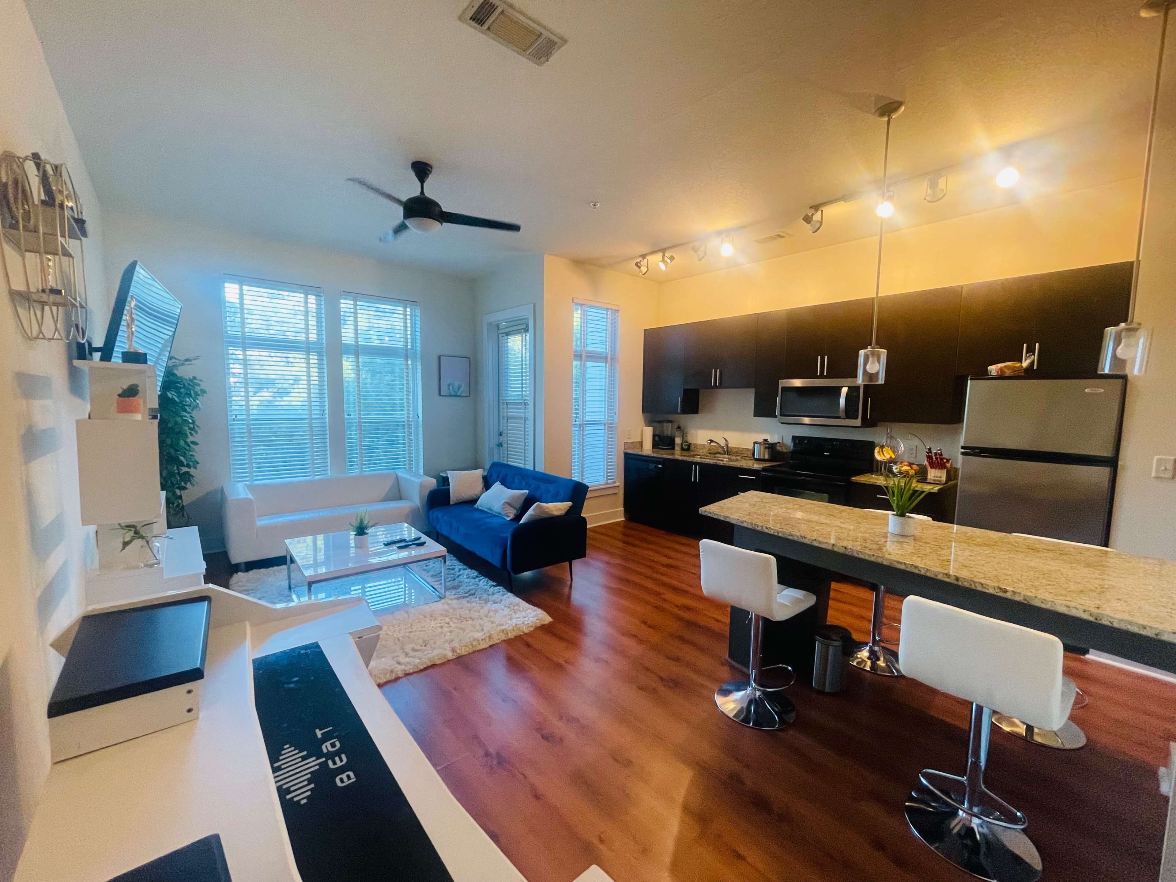 A modern apartment with a living area furnished with a white sofa, blue couch, and a kitchen featuring dark cabinets and a granite countertop.