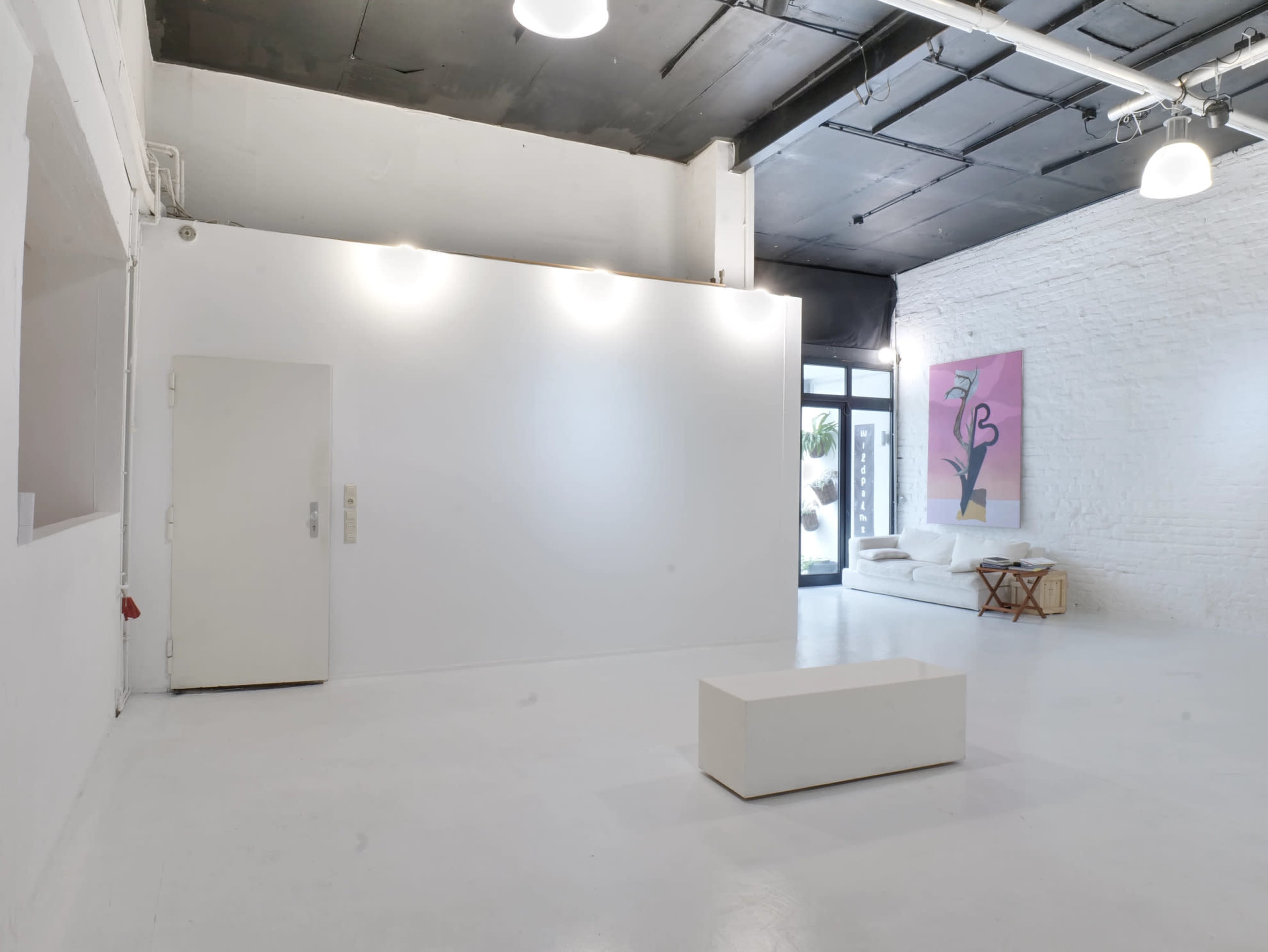 The image shows a minimalist art gallery with white walls, a simple white bench in the center, and a colorful artwork displayed on one wall.