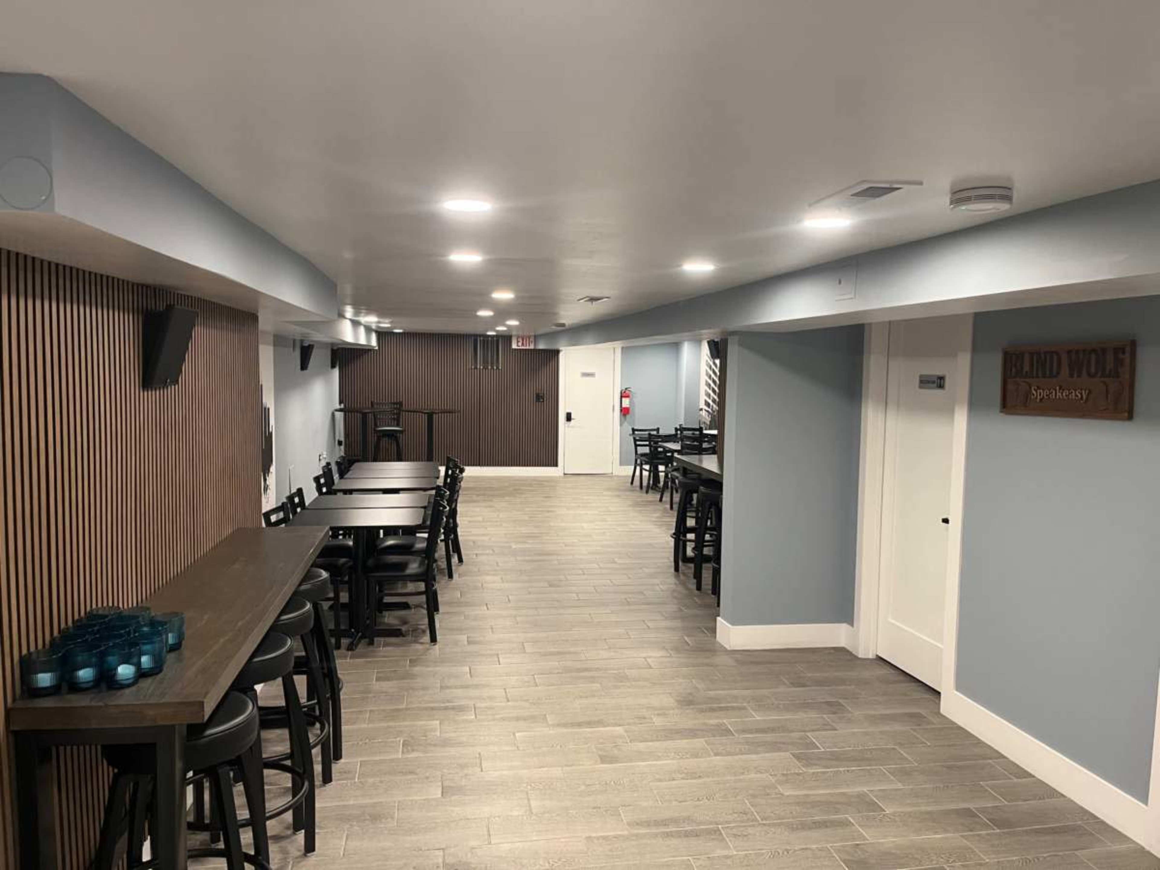 The image shows a narrow, well-lit hallway with tables and chairs on either side, leading to a door at the end and a sign on the wall indicating "Blind Wolf Speakeasy."