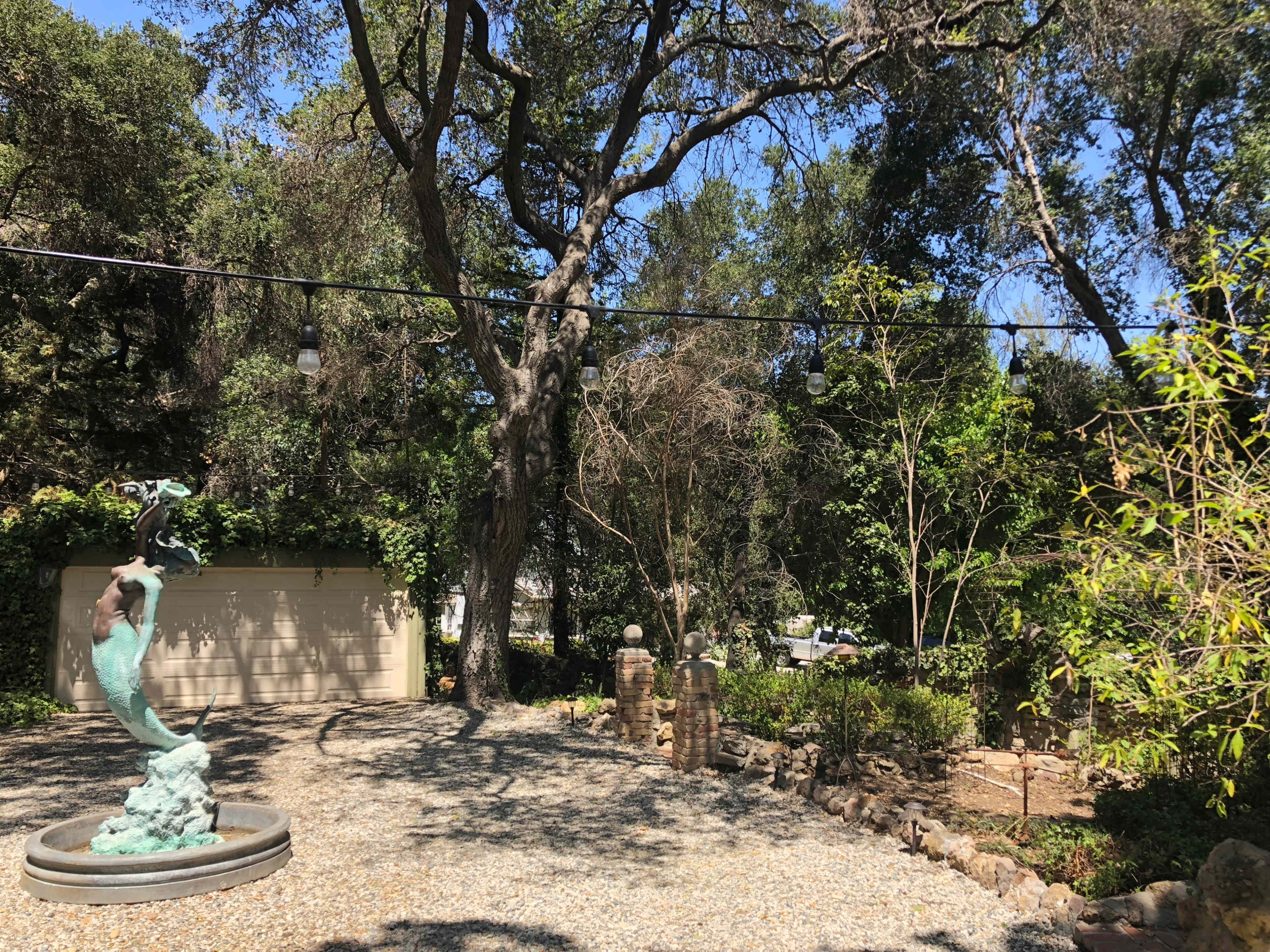 A sculpture stands in a gravel area surrounded by trees and gardens under a clear blue sky.