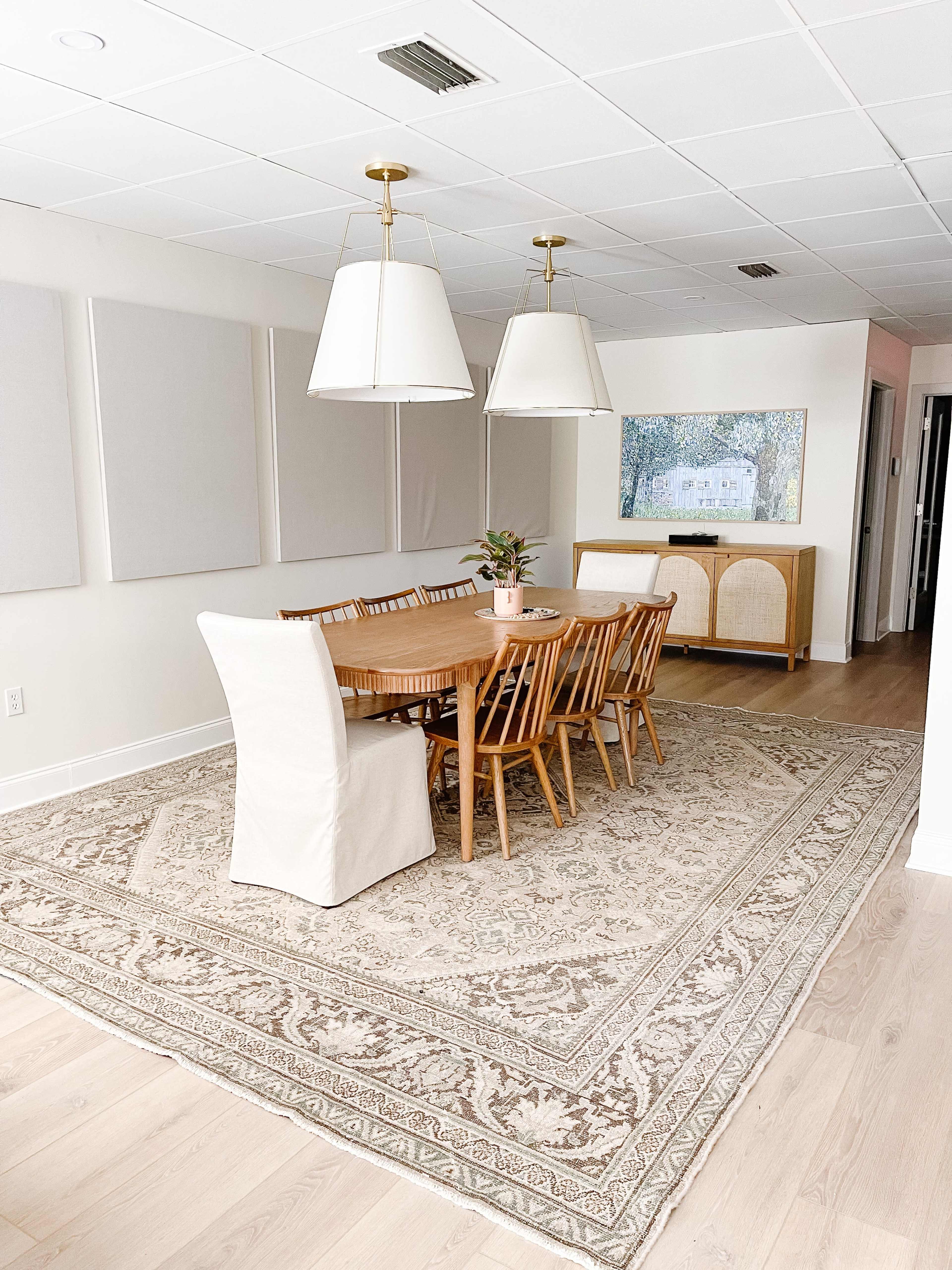 A dining room features a wooden table surrounded by eight chairs on a patterned rug, with two pendant lights hanging above and a sideboard along the wall.