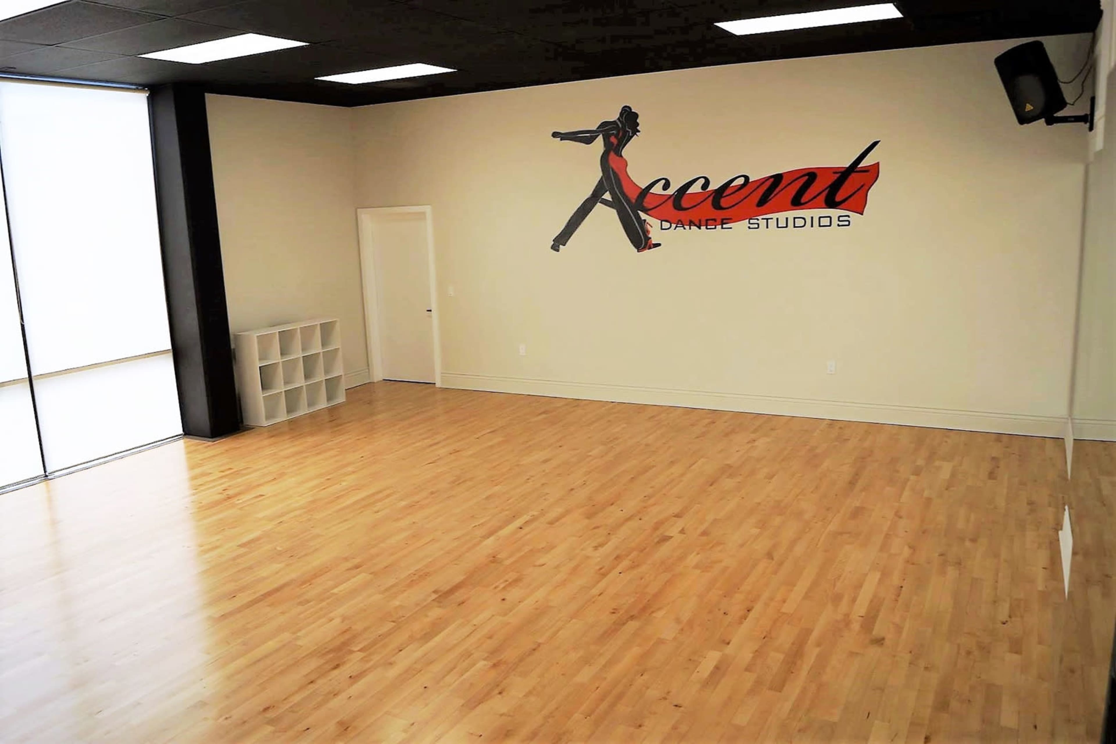 An empty dance studio with wooden floors, a large window, and a wall featuring a sign that reads "Accent Dance Studios."