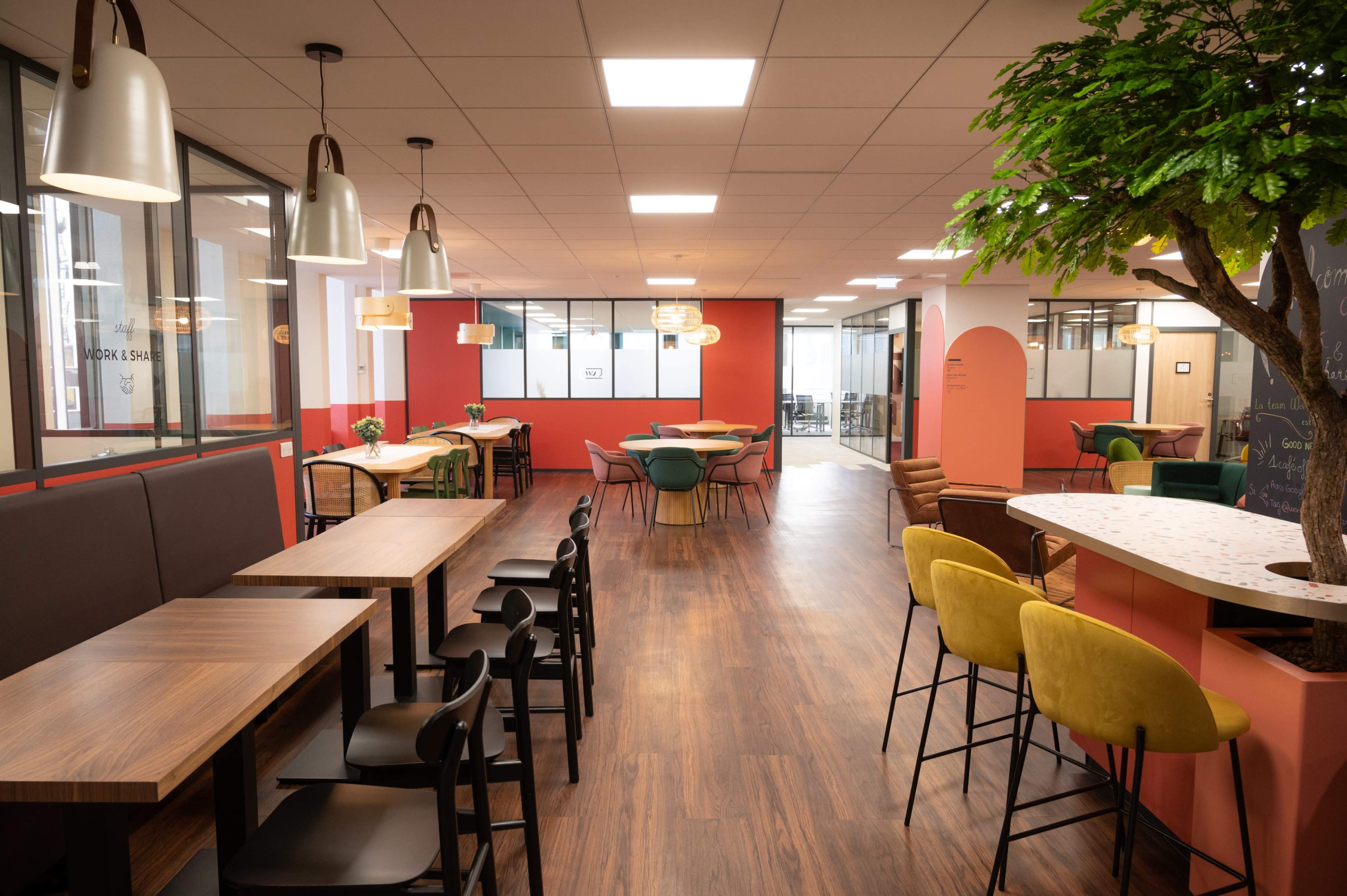A modern office space with various seating arrangements, including wooden tables and colorful chairs, along with large windows and plants.