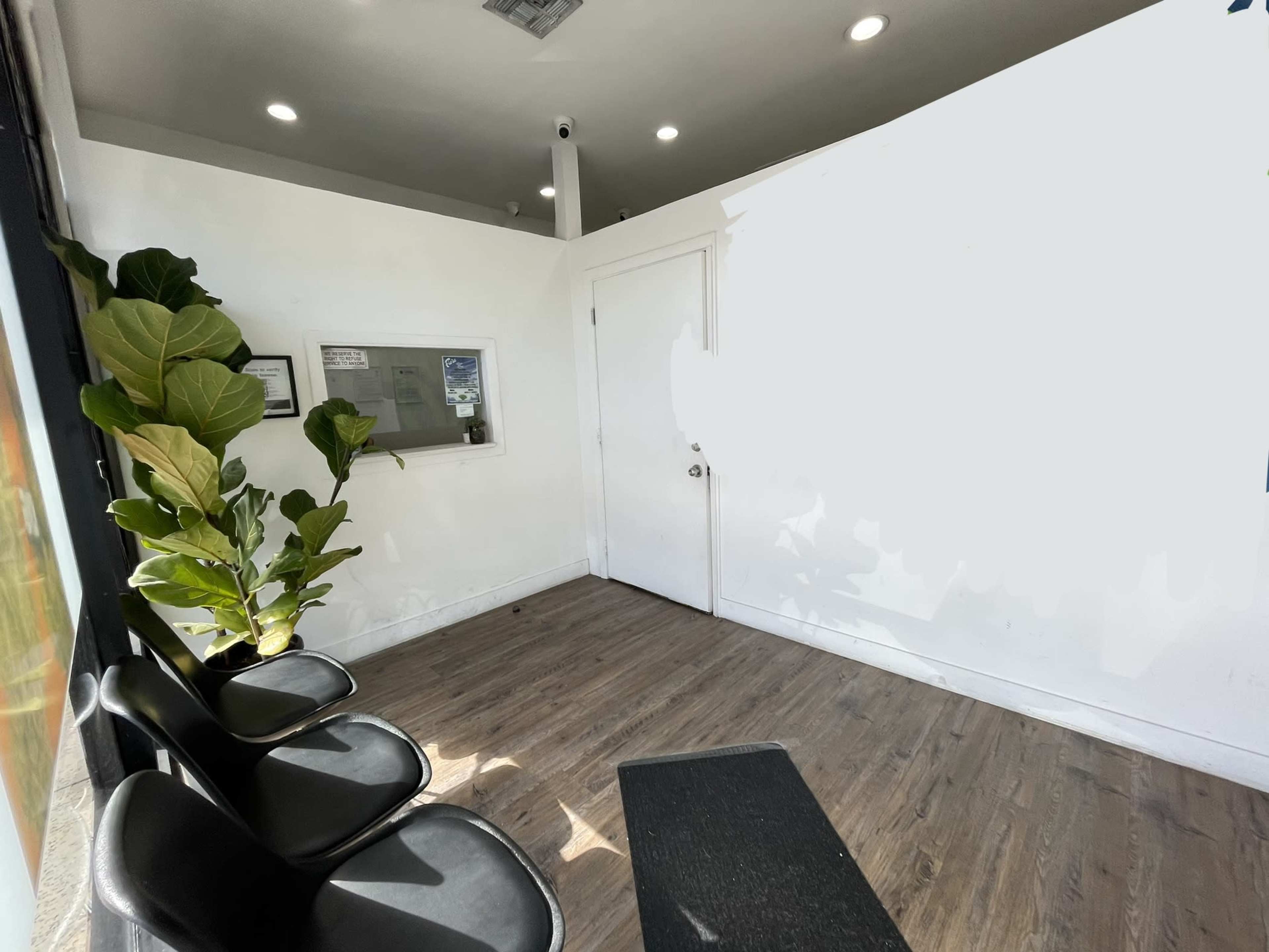 The image shows a minimalistic waiting area with three black chairs, a small plant, and a door leading to another room.