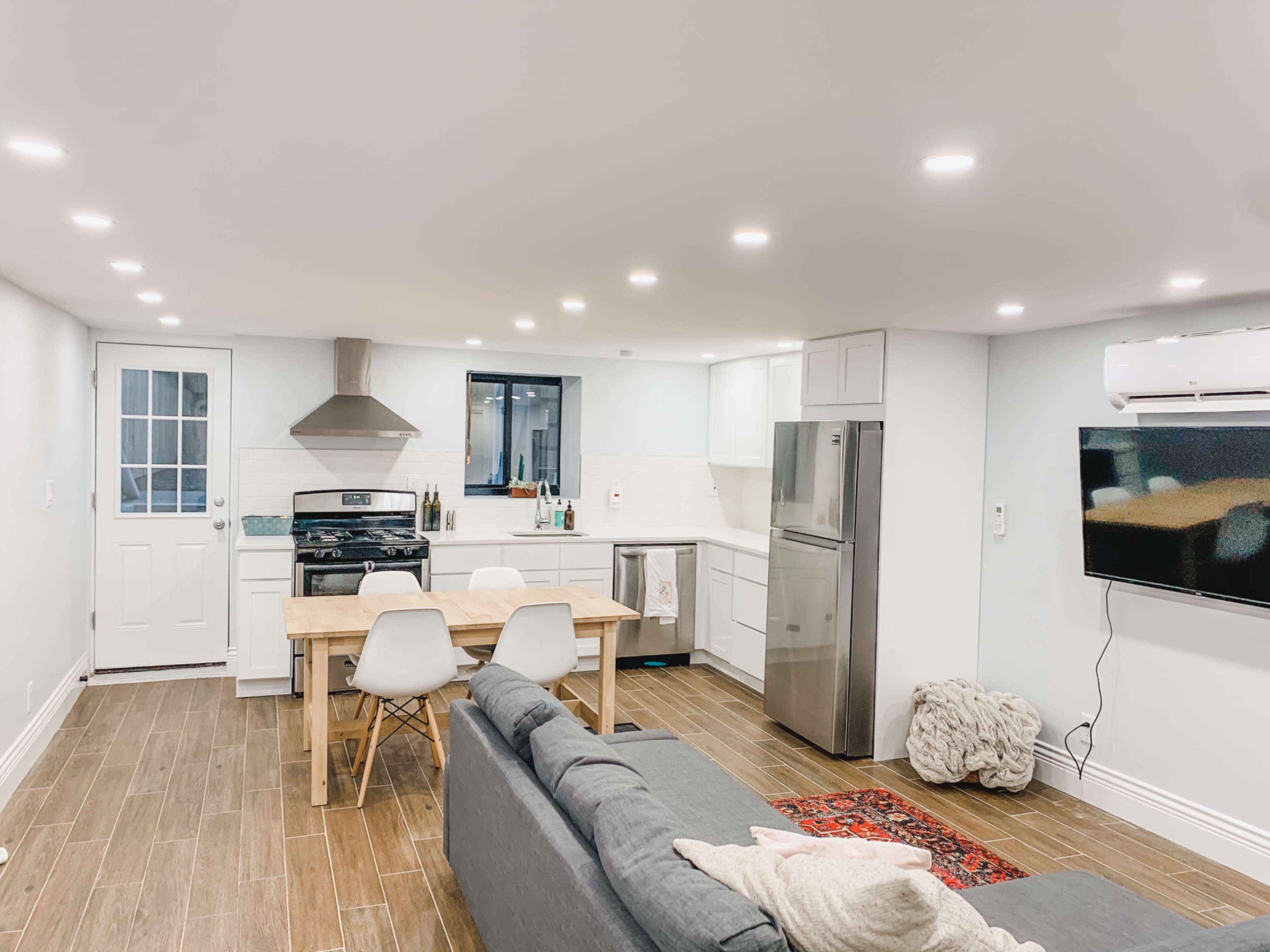 A modern kitchen with a dining table and chairs, a gray sofa, and a flat-screen TV in a well-lit space.