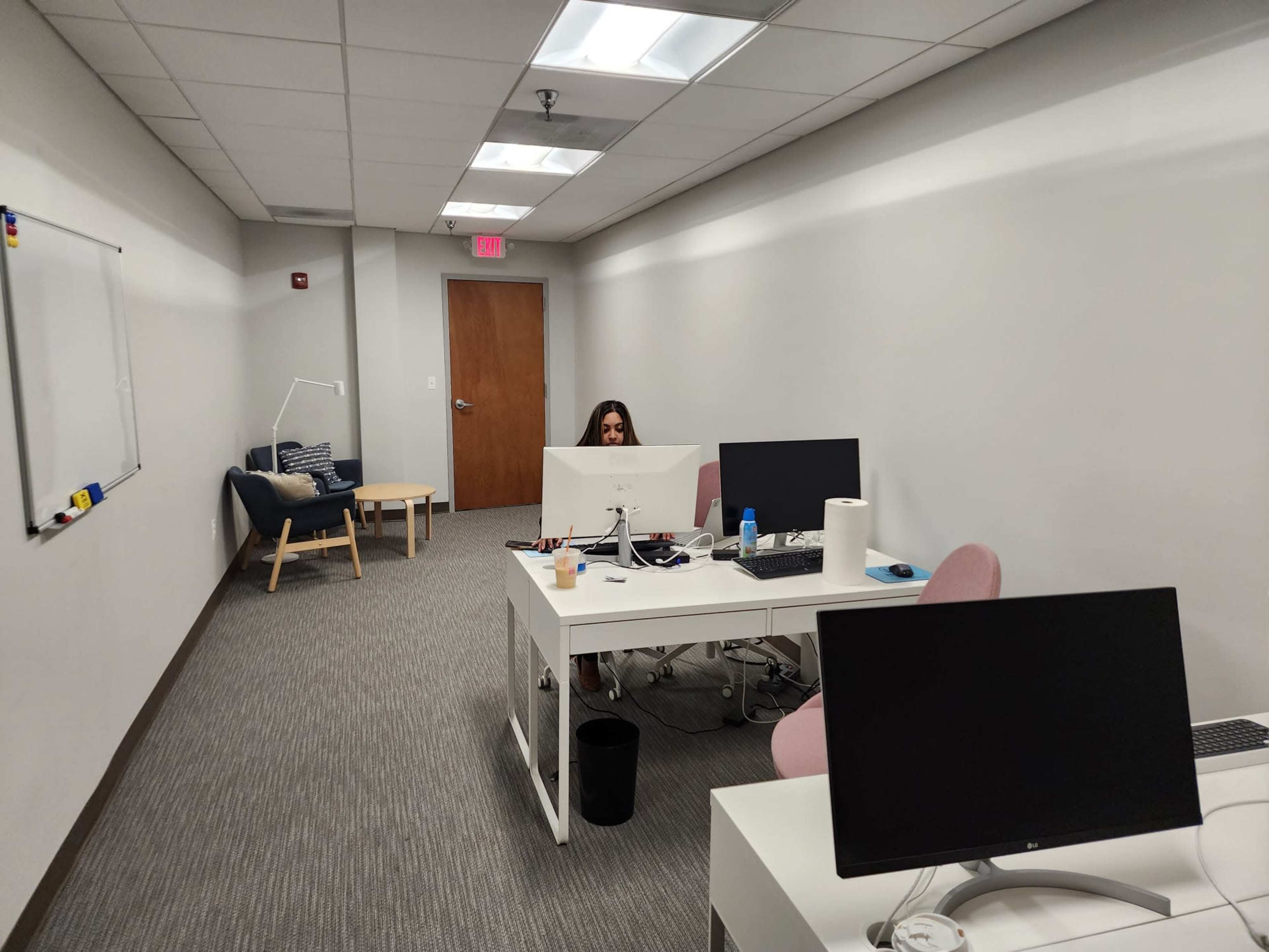 A person sits at a desk in a minimalist office space with two computer monitors, while a seating area with a chair and floor lamp is visible in the background.