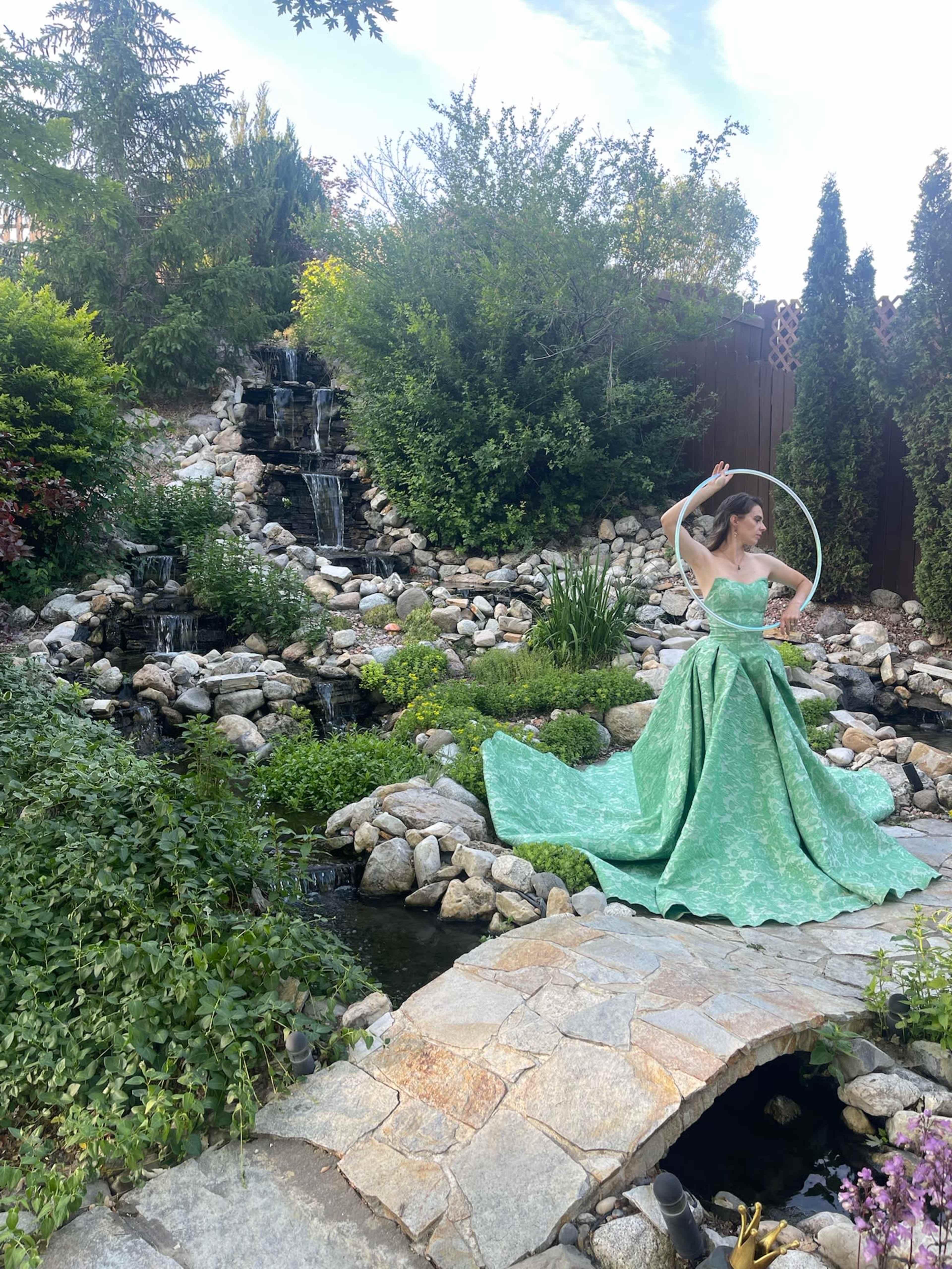 A woman in a green gown poses with a hoop in a rock-lined garden featuring a small waterfall and lush vegetation.