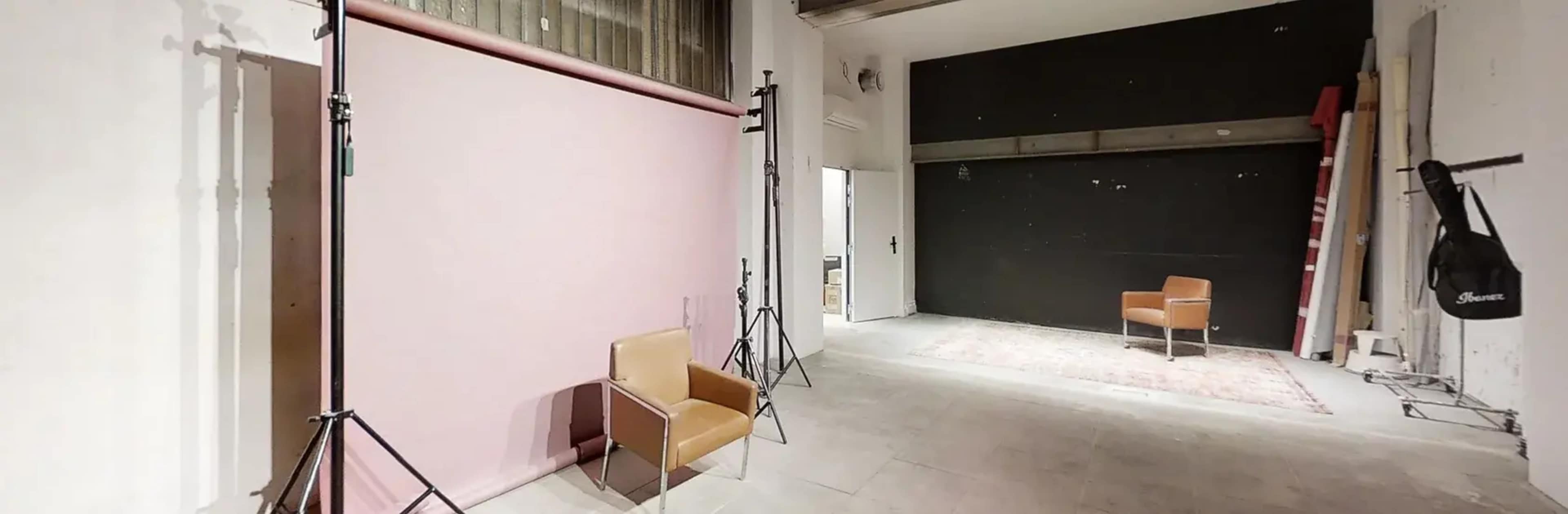 The image shows a photography studio with a pink backdrop, two light stands, a brown chair, and a black wall.