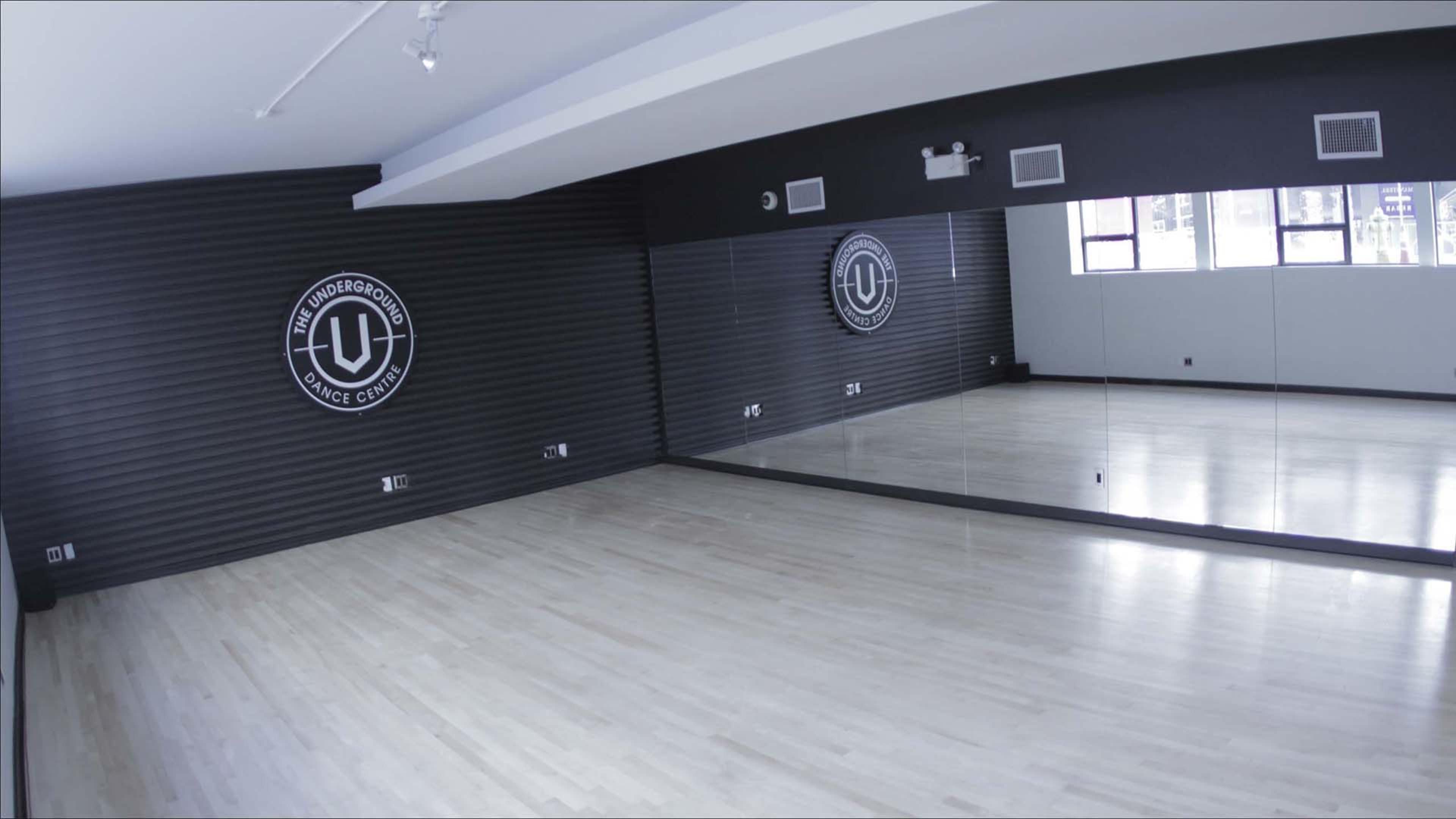 A spacious dance studio with hardwood flooring, large mirrors, and a logo displayed on the wall.