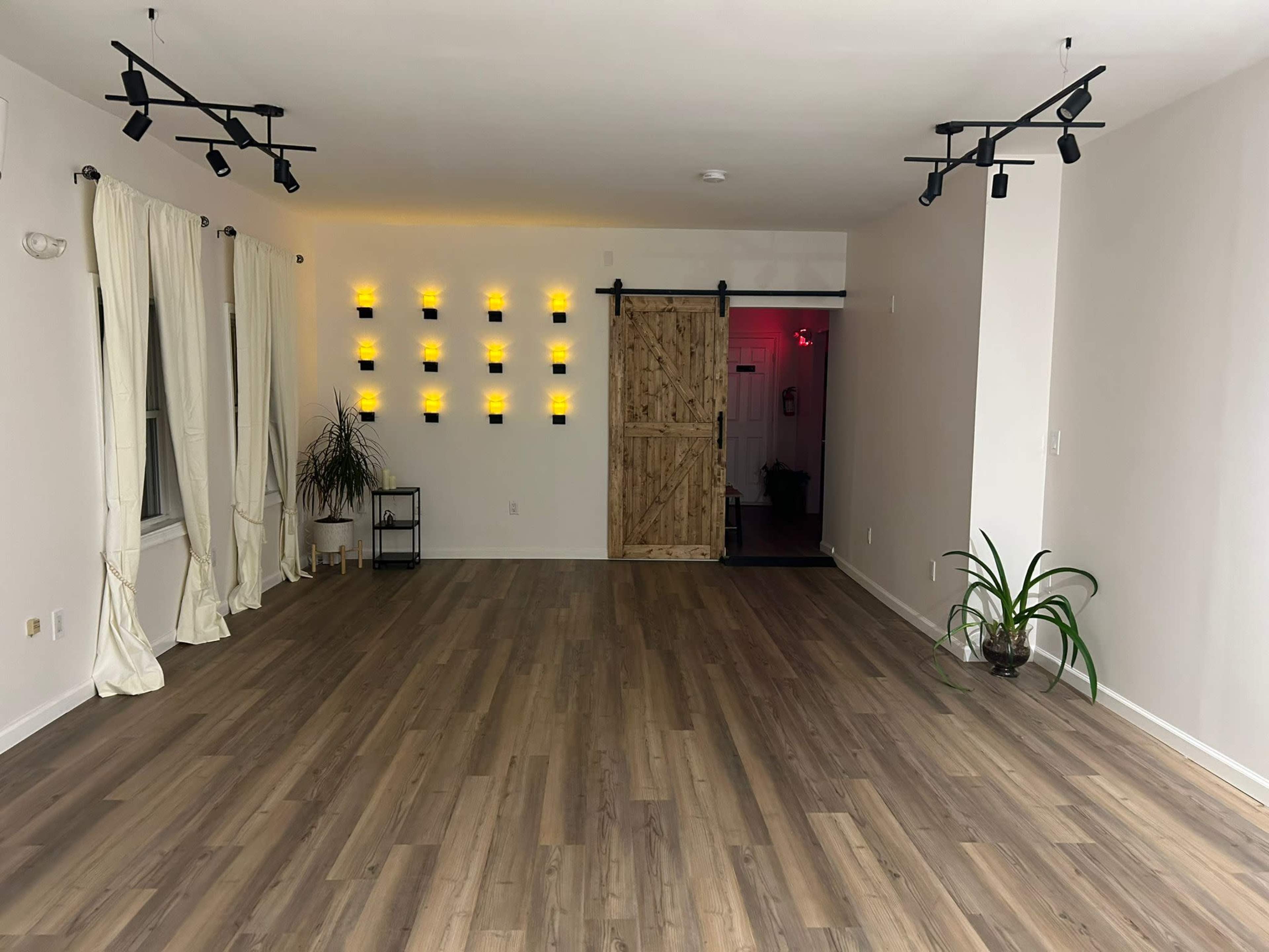 A spacious, empty room with wooden flooring, a barn-style sliding door, track lighting, and a wall adorned with yellow light fixtures.