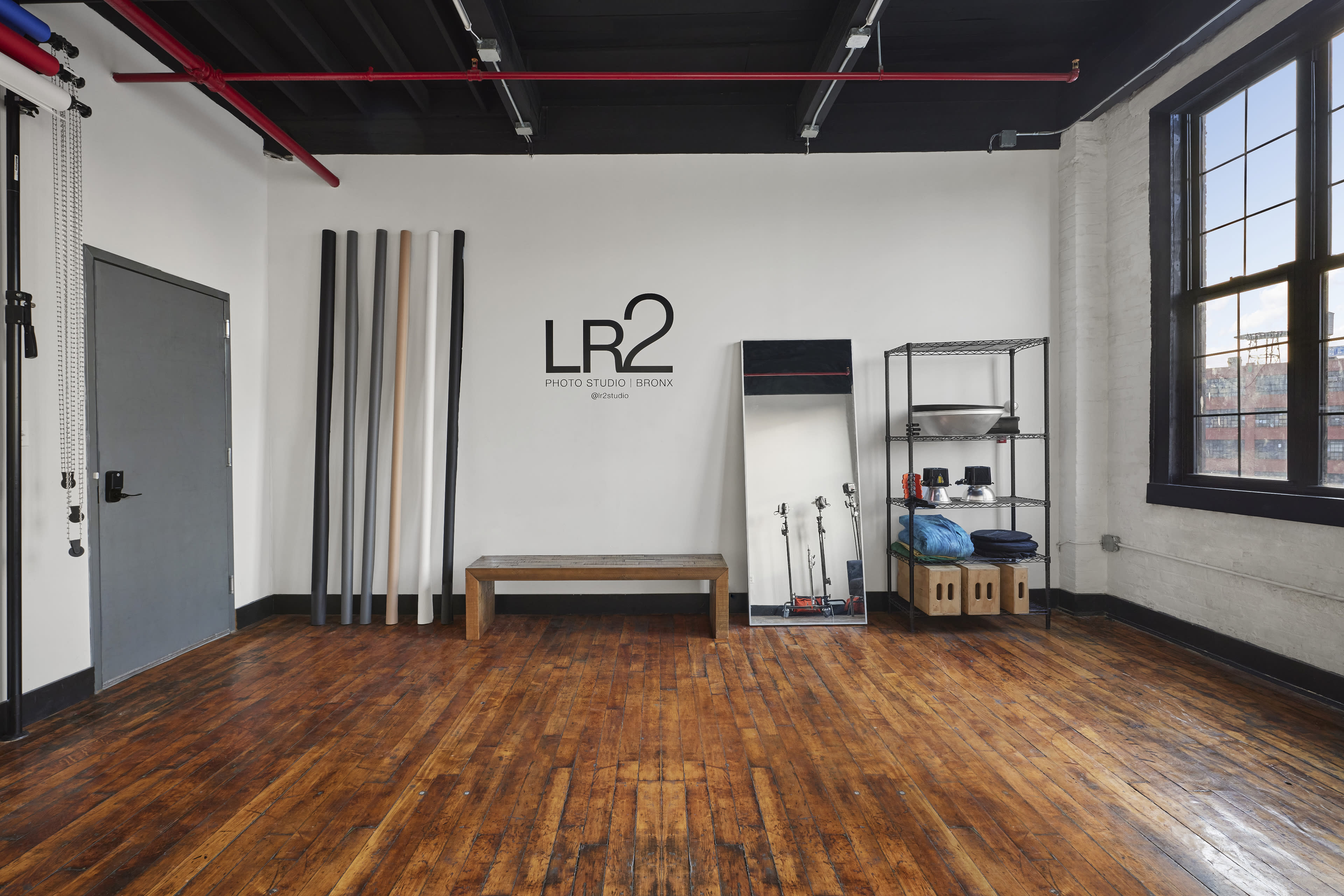 A spacious studio featuring wooden floors, a mirror, a bench, and several large poles against a white wall with the text "LR2 Photo Studio Brooklyn."