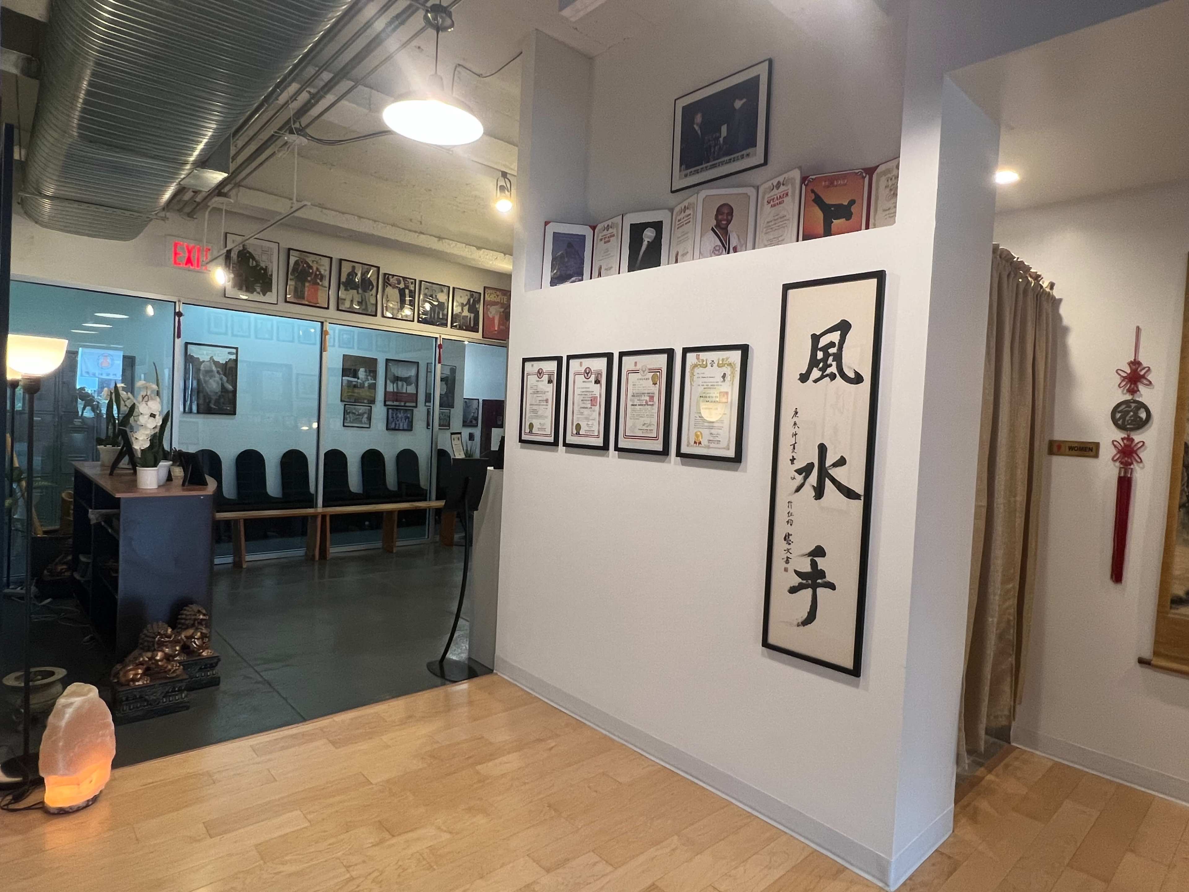 A hallway with framed certificates and artwork on the walls, leading to a waiting area with chairs and a glass partition.