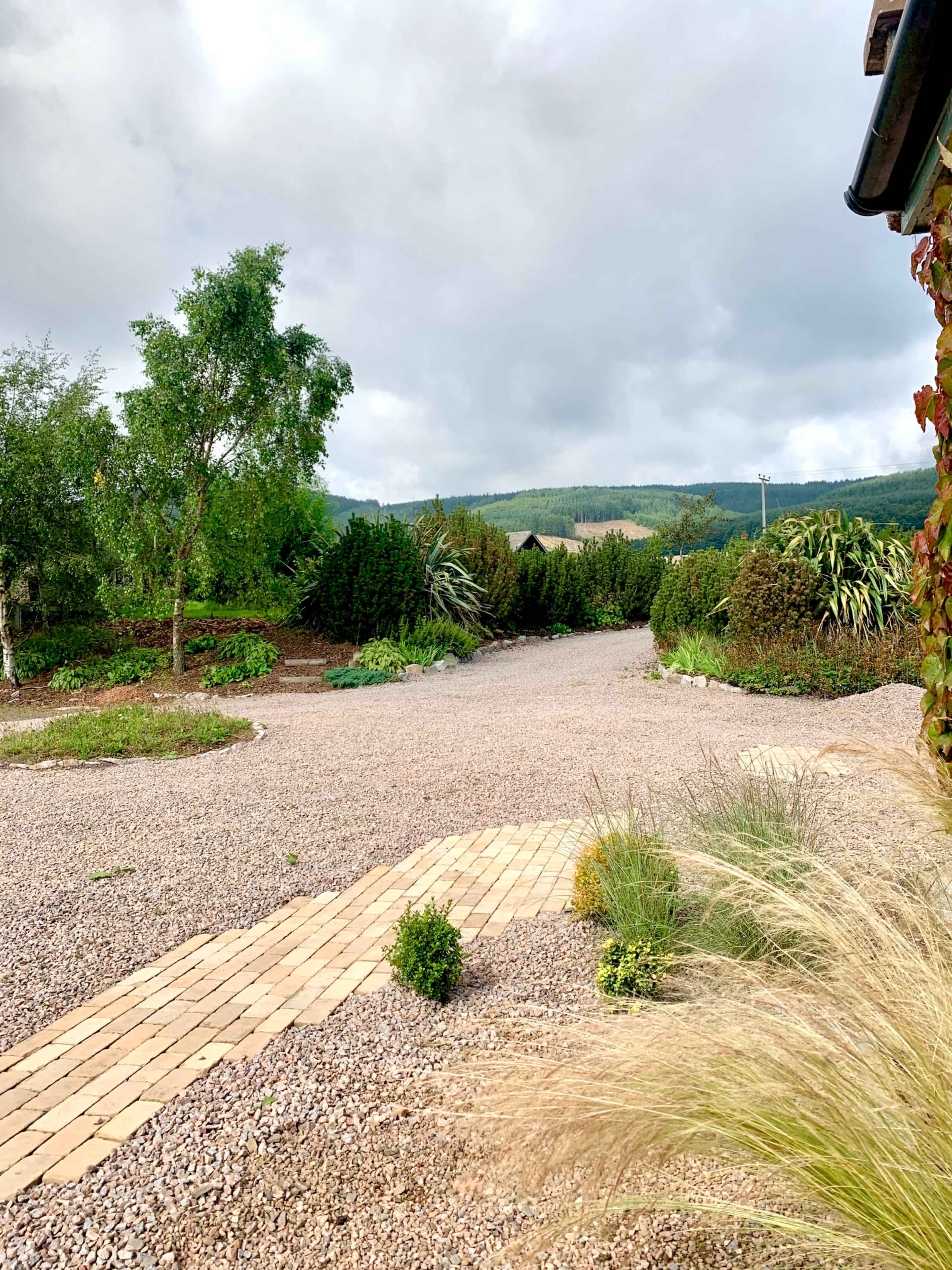 A gravel pathway leads through a landscaped area with various plants and trees, set against a cloudy sky and distant hills.