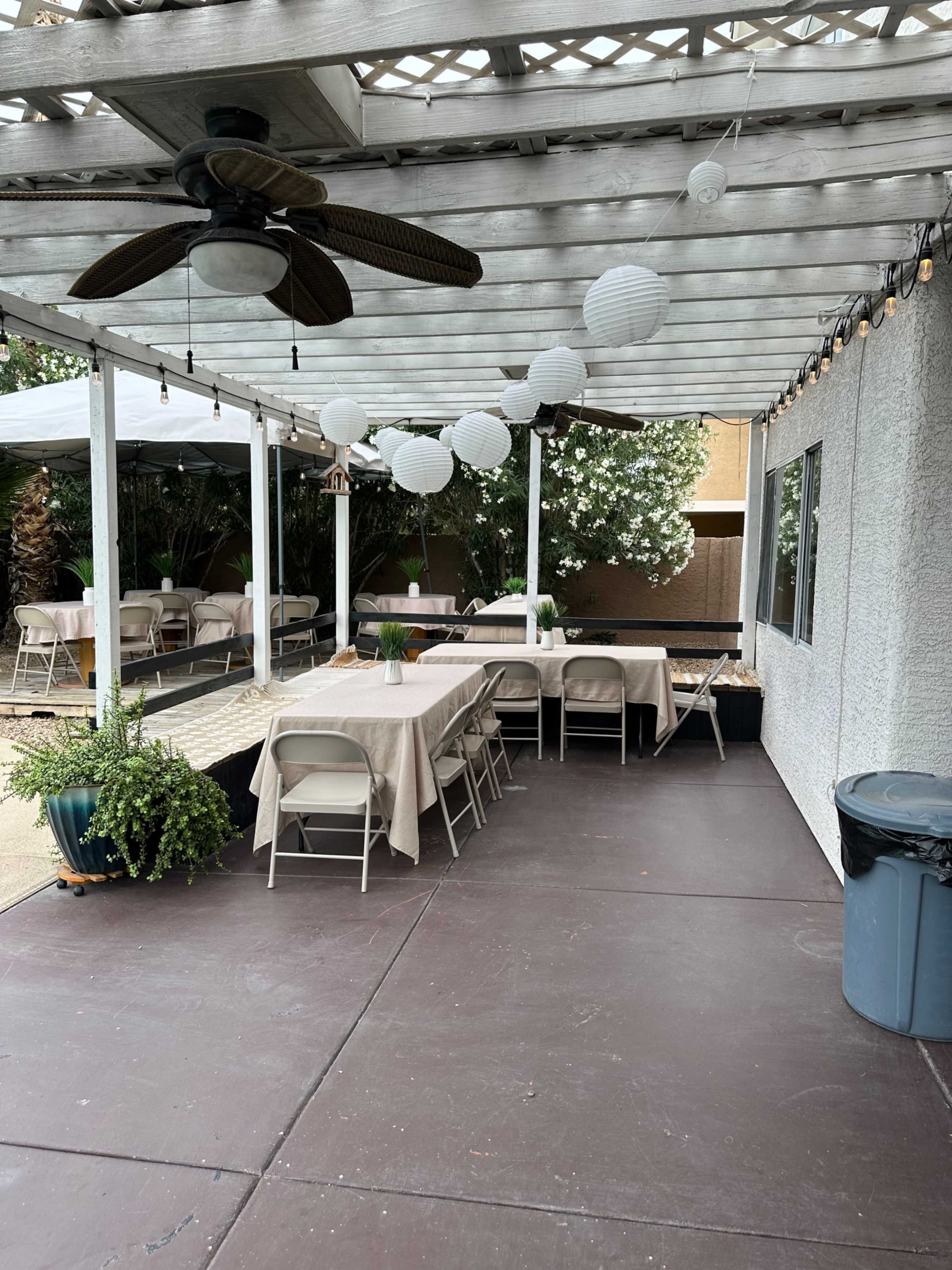 A covered outdoor patio features several tables set for an event, with decorative paper lanterns hanging above and potted plants nearby.
