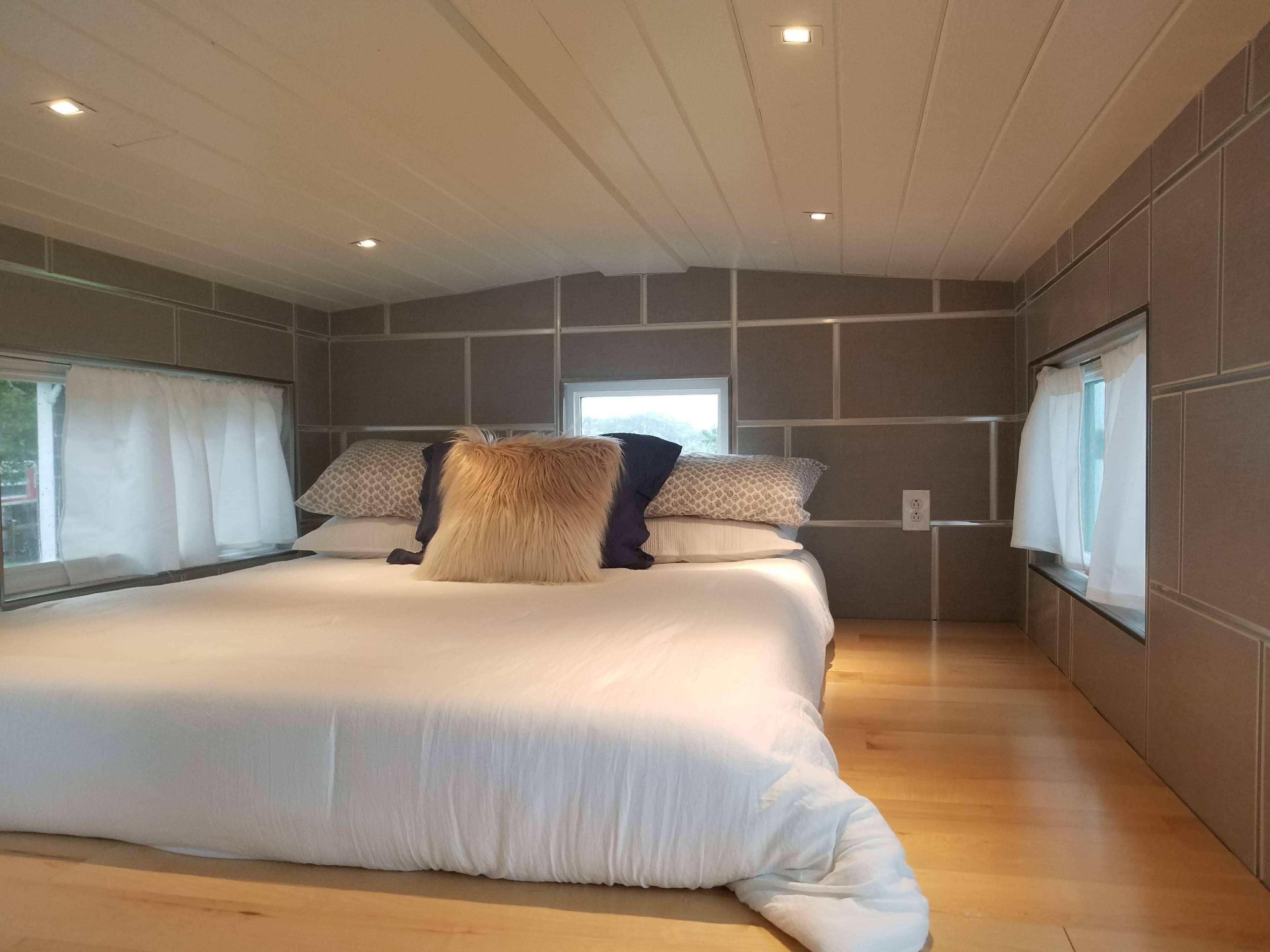 A cozy bedroom with a large white bed adorned with decorative pillows, set against gray paneled walls and framed by windows.