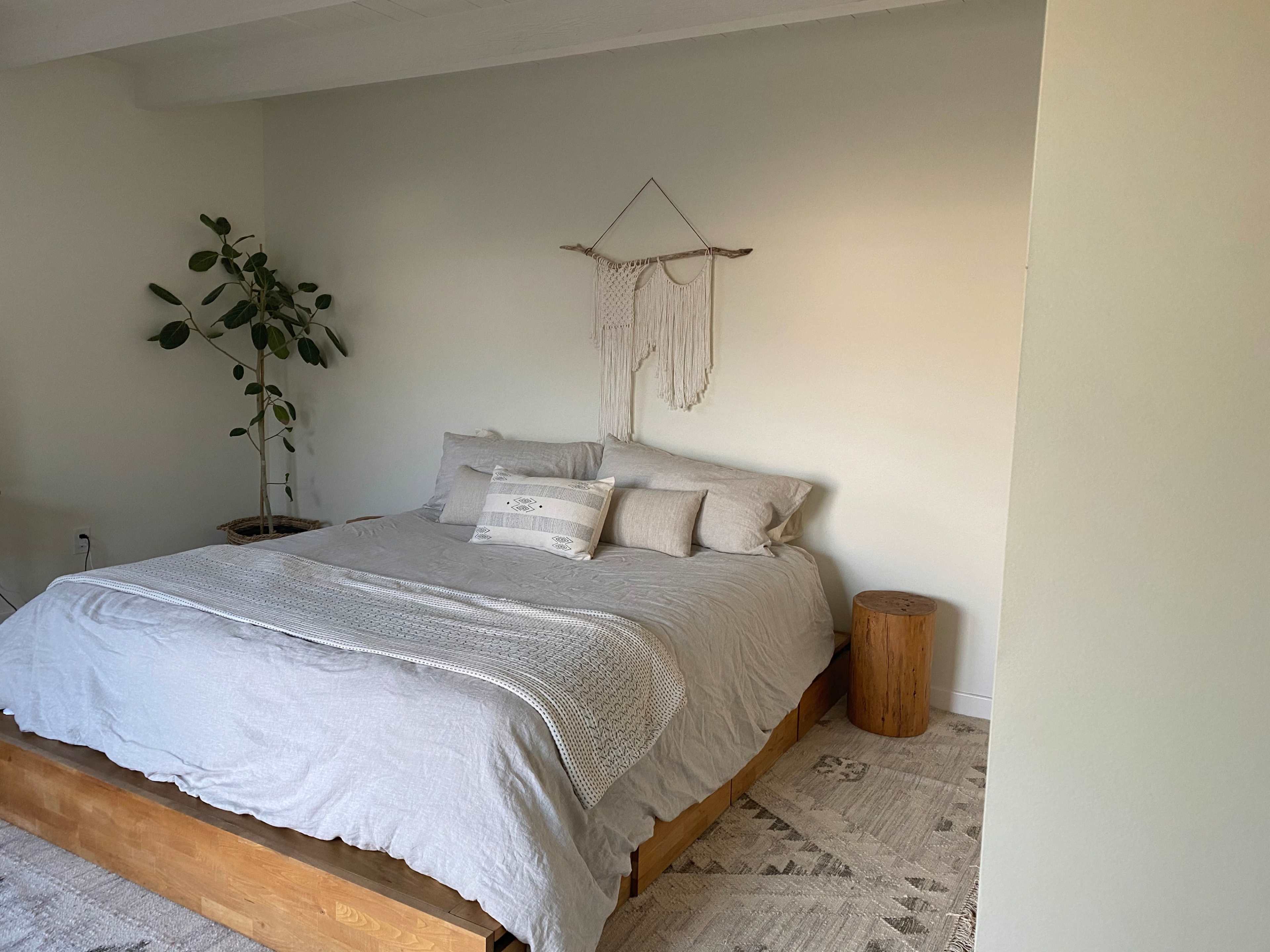 The image shows a bedroom with a large bed covered in light gray bedding, a decorative wall hanging above it, a potted plant in the corner, and a wooden stool beside the bed.
