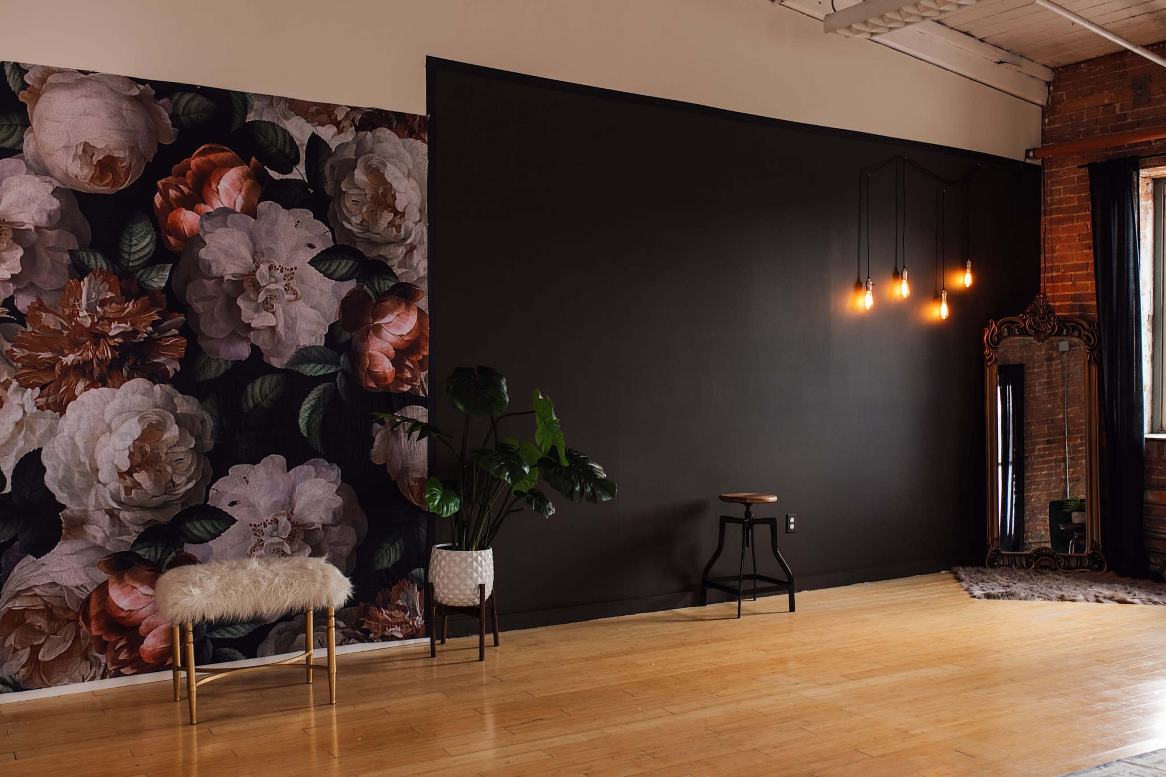 The image shows a spacious room with a floral wallpaper on one side, a dark wall, a vintage mirror, a stool, and plants, illuminated by industrial-style pendant lights.