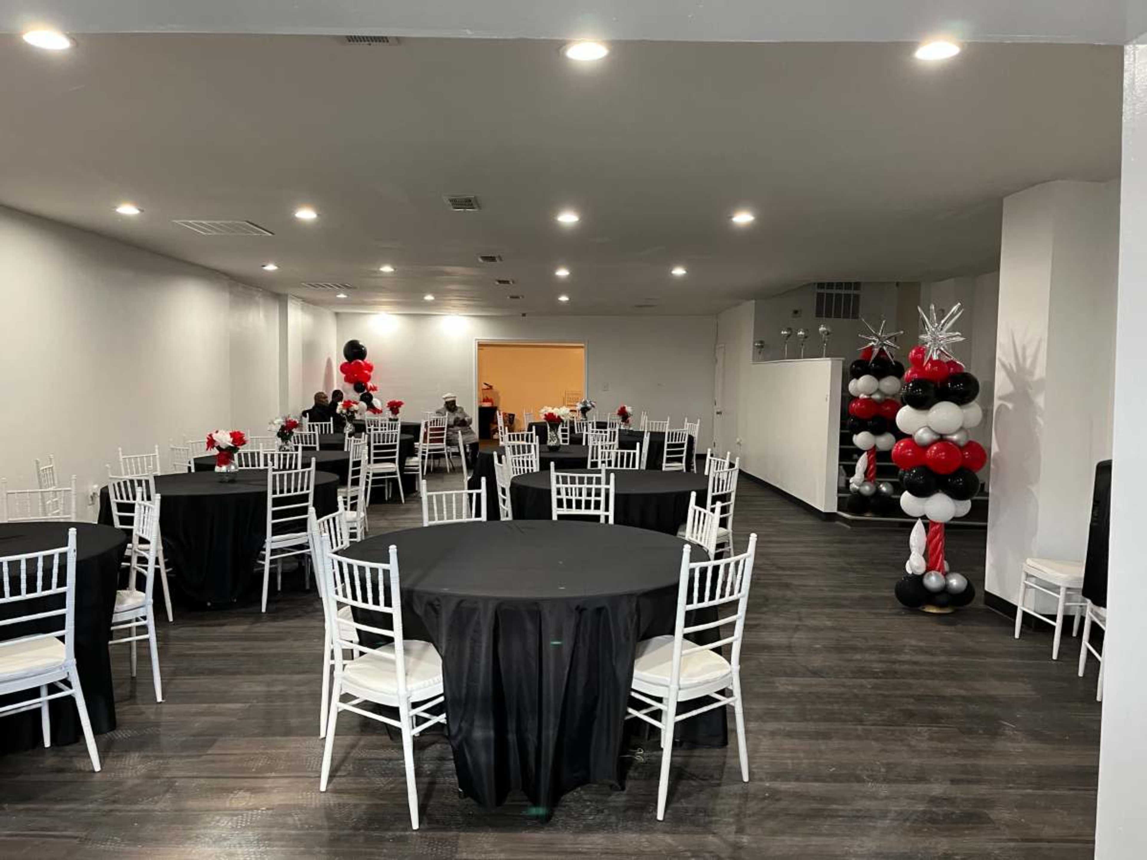 A spacious event room features several tables adorned with black tablecloths, white chairs, and decorative balloon arrangements in red, black, and white.
