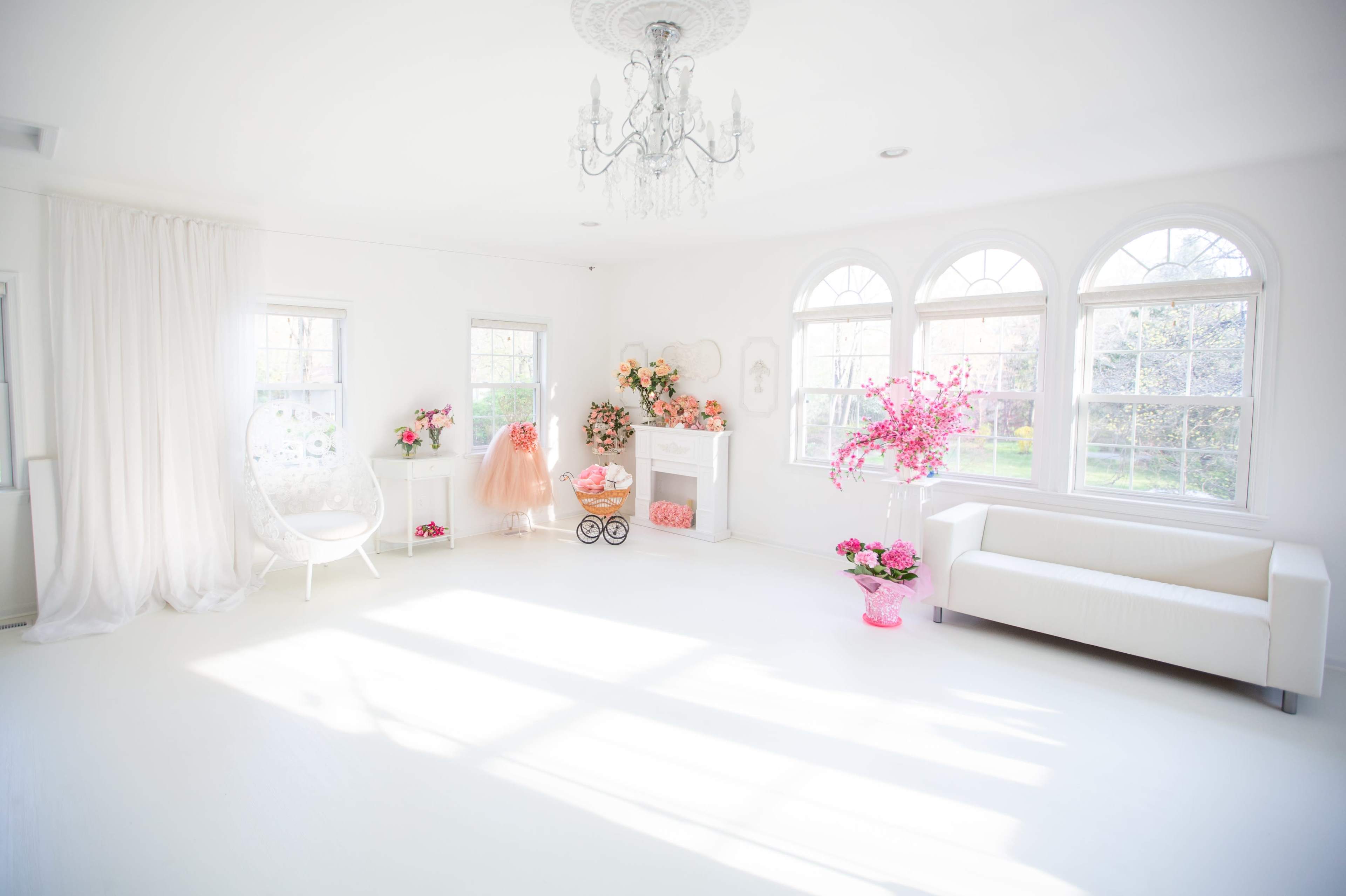 The image shows a bright room with large windows, a white sofa, a hanging chair, and pink decorative elements, including flowers and a dress.