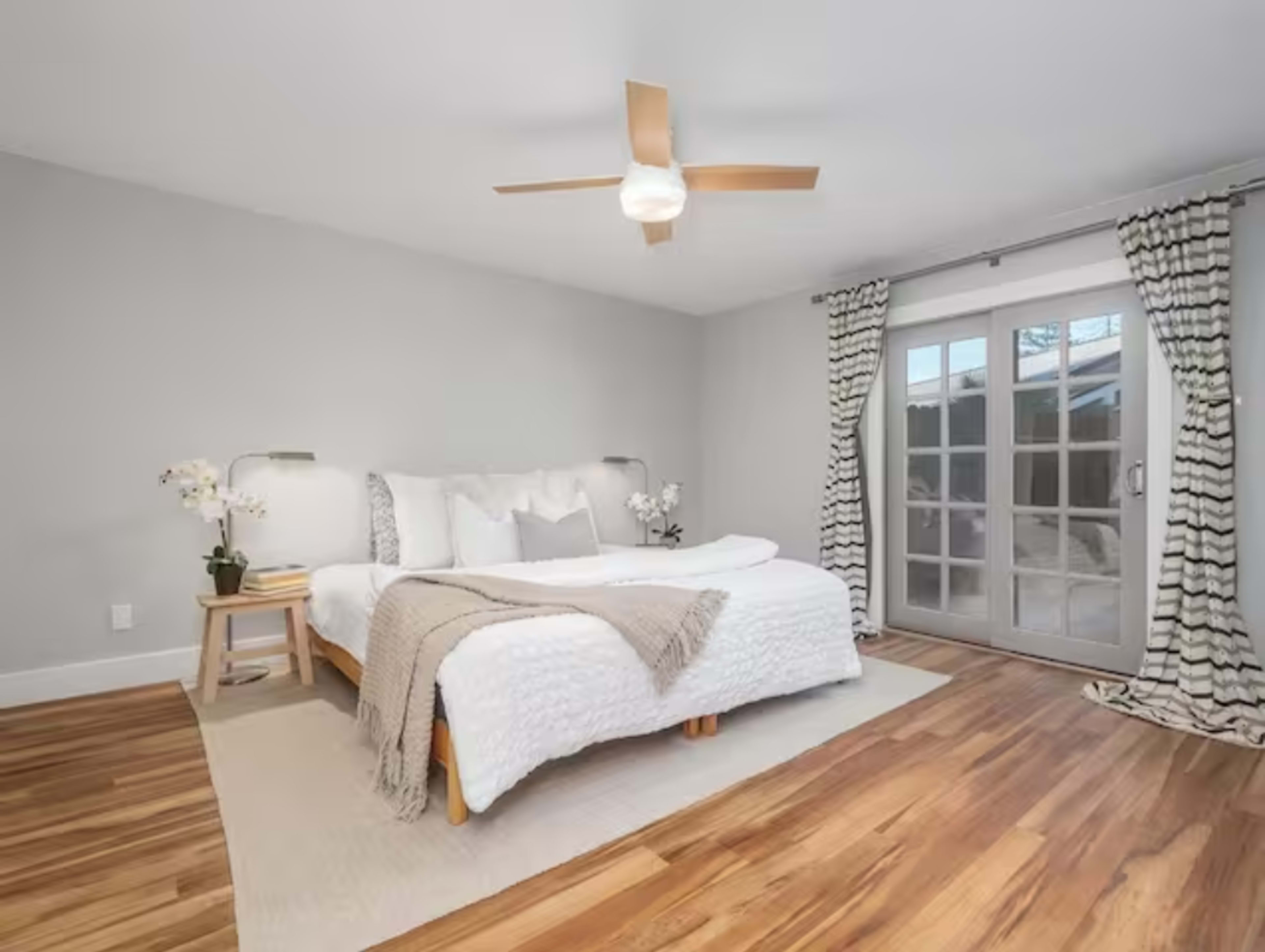 A modern bedroom featuring a bed with a gray throw blanket, a ceiling fan, wood flooring, and a set of glass patio doors leading to an outdoor space.
