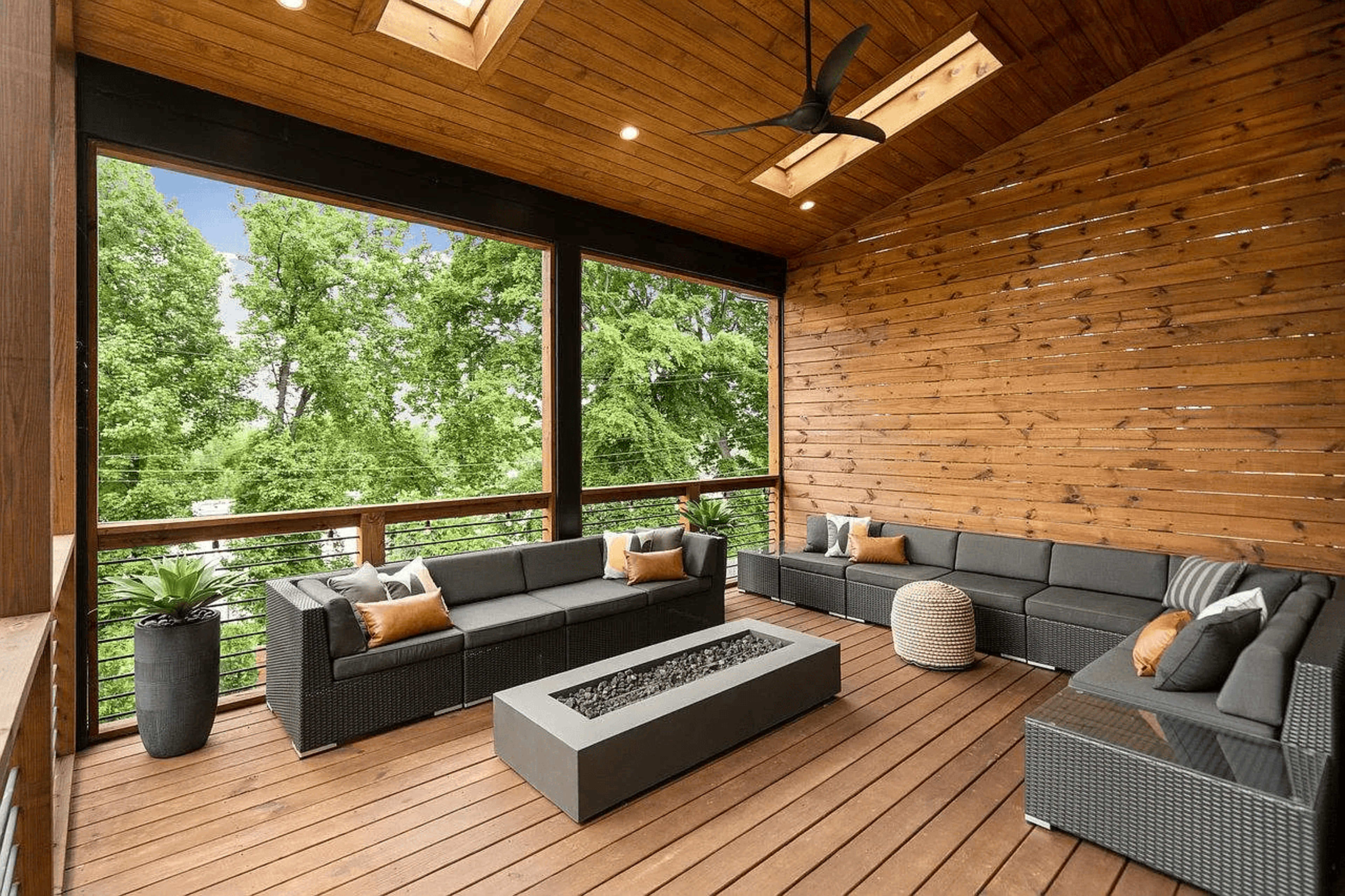 A spacious covered patio features modern seating arrangements and a fire pit, surrounded by wooden walls and large windows that open to a view of trees.