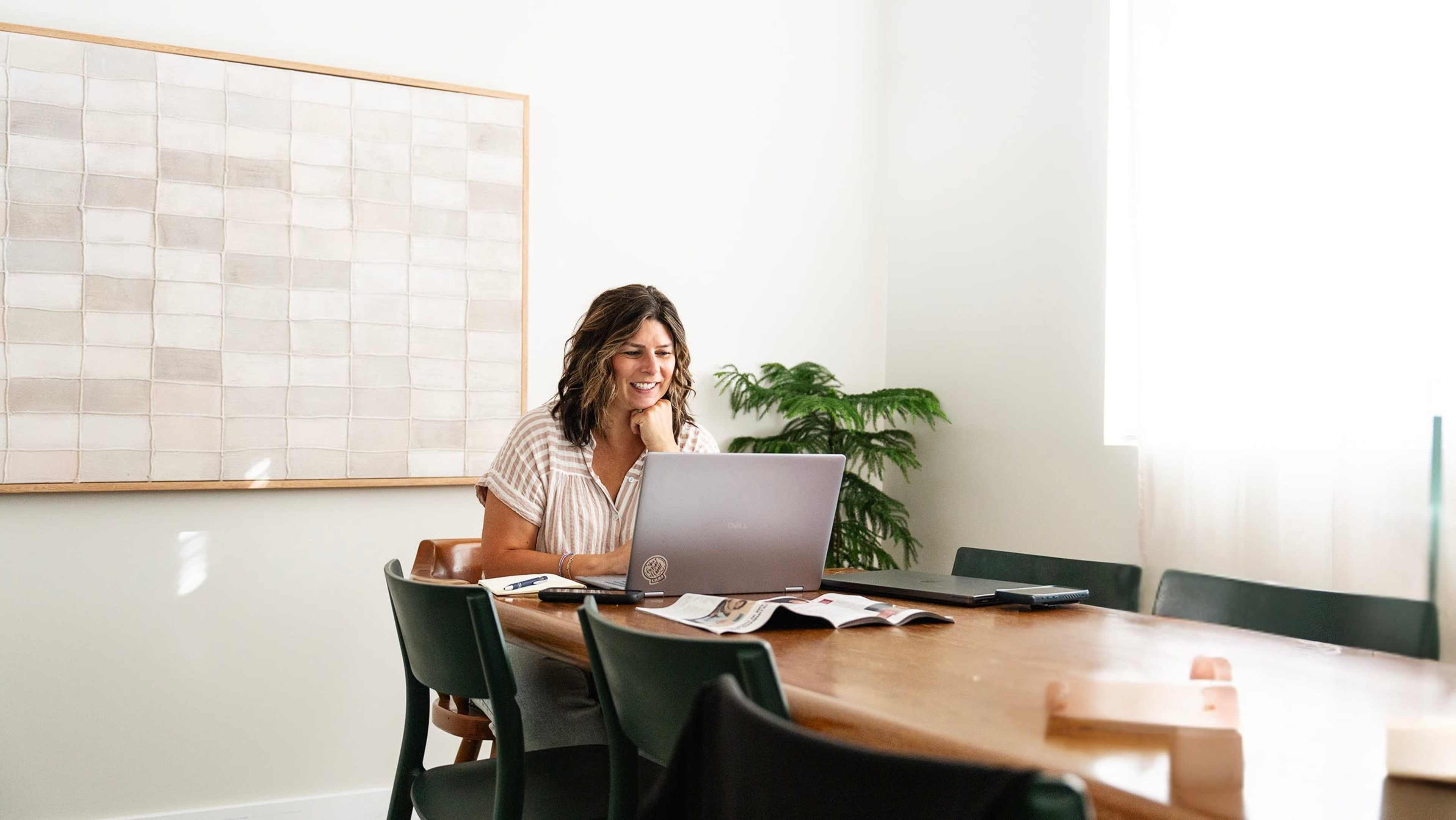 A woman with wavy hair sits at a wooden table with a laptop, smiling as she works in a well-lit room featuring a large art piece on the wall and a potted plant nearby.