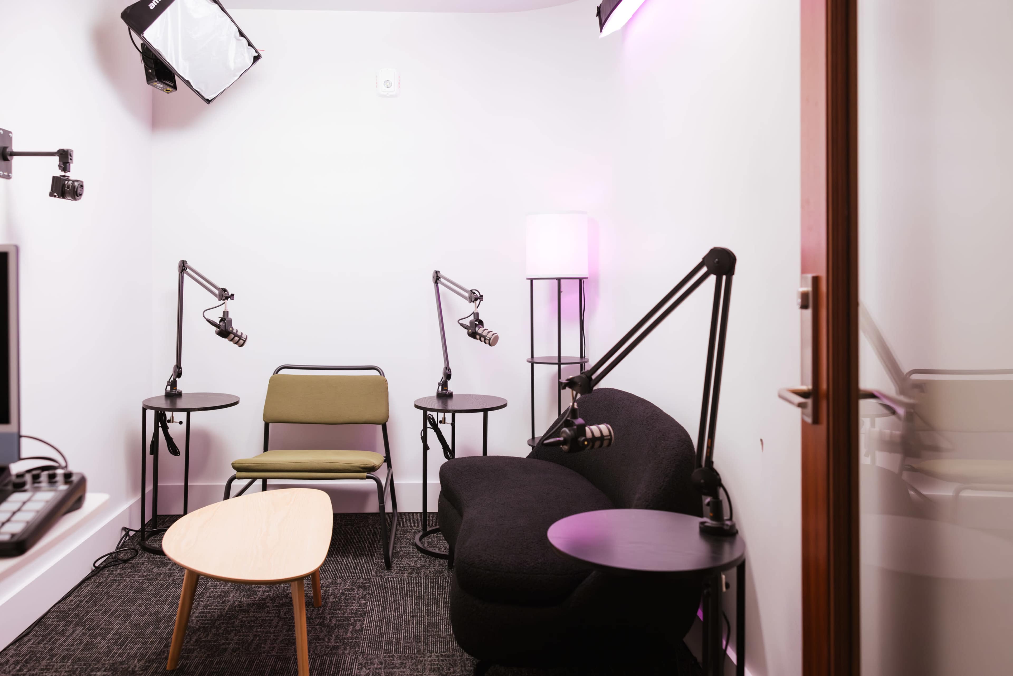 The image shows a small, modern recording studio with two audio equipment stands, a sofa, a chair, and a round coffee table.