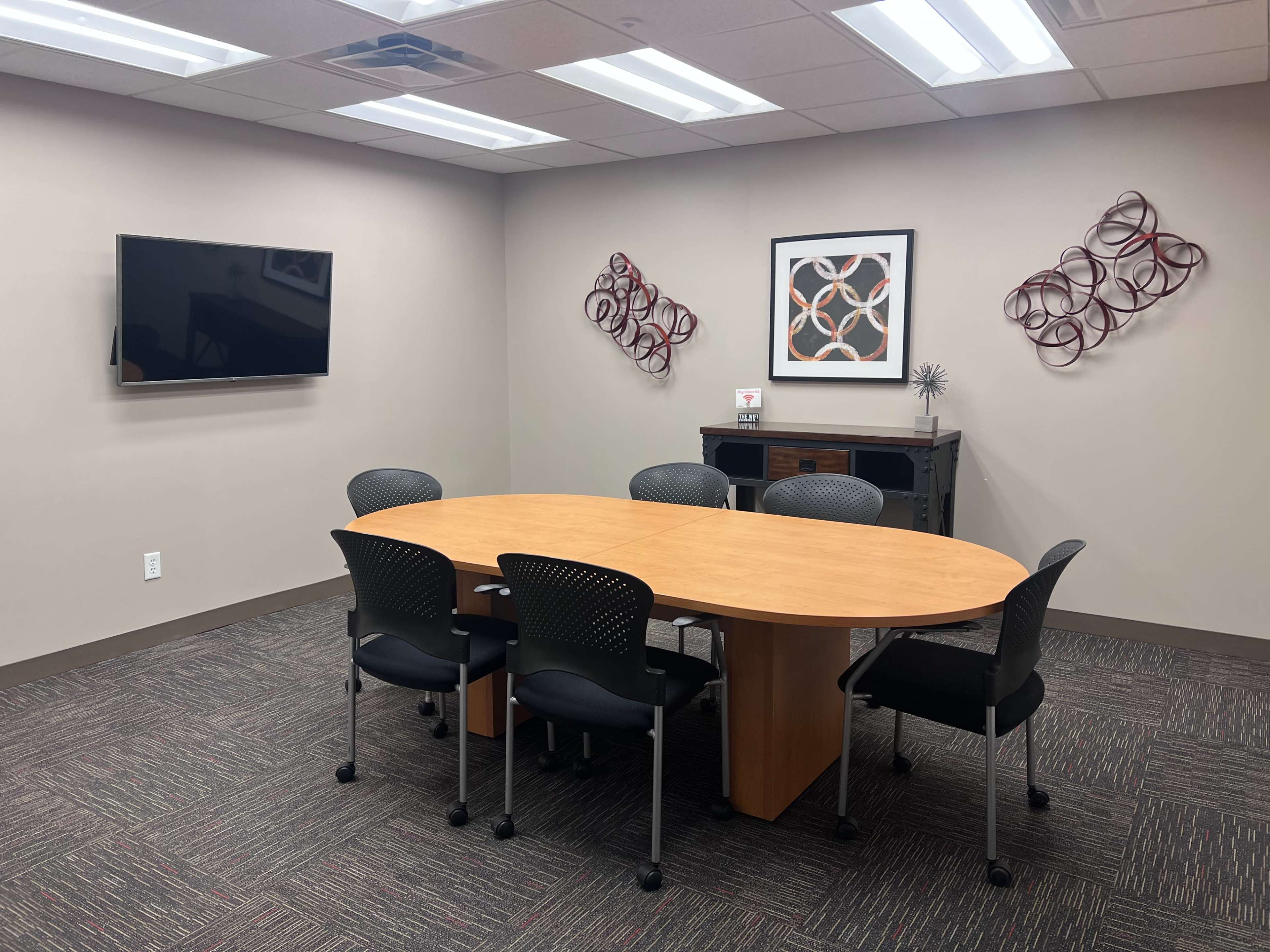The image shows a conference room with a large oval wooden table surrounded by six black chairs, a wall-mounted television, and decorative wall art.