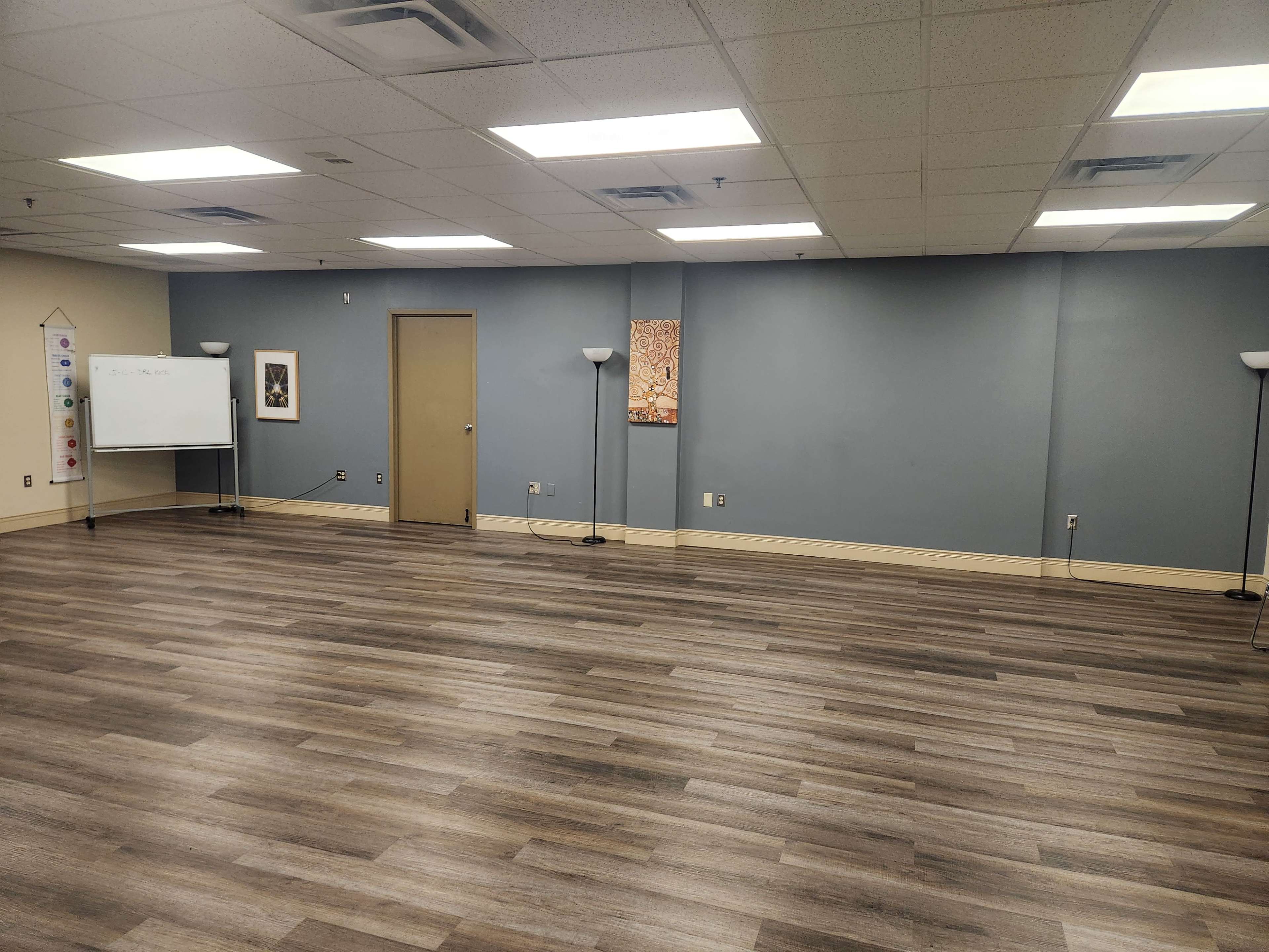 A spacious, empty room with gray walls, wooden flooring, a whiteboard, and floor lamps.