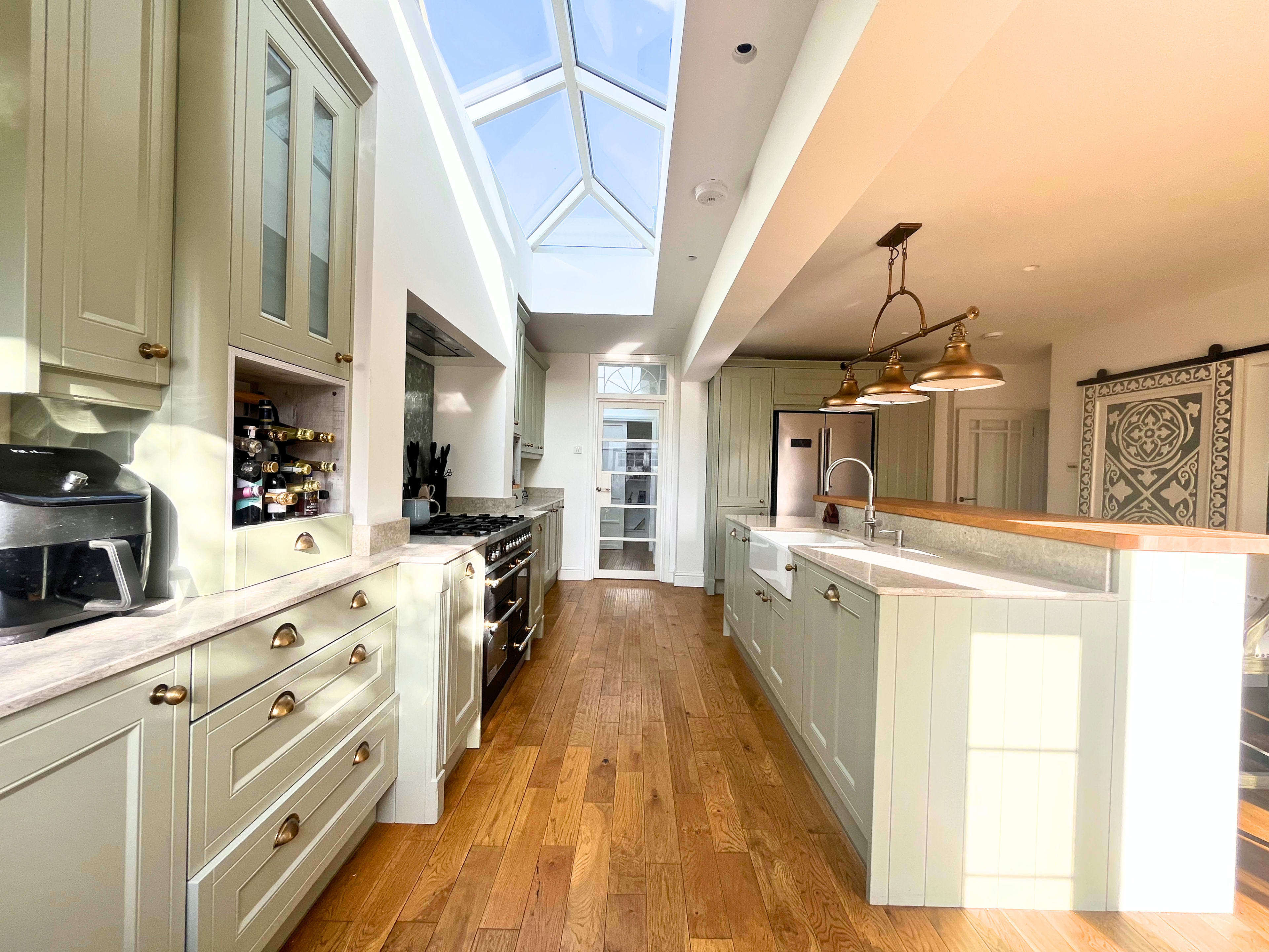 The image shows a modern kitchen with light green cabinetry, a large skylight, wooden flooring, and a spacious open layout.