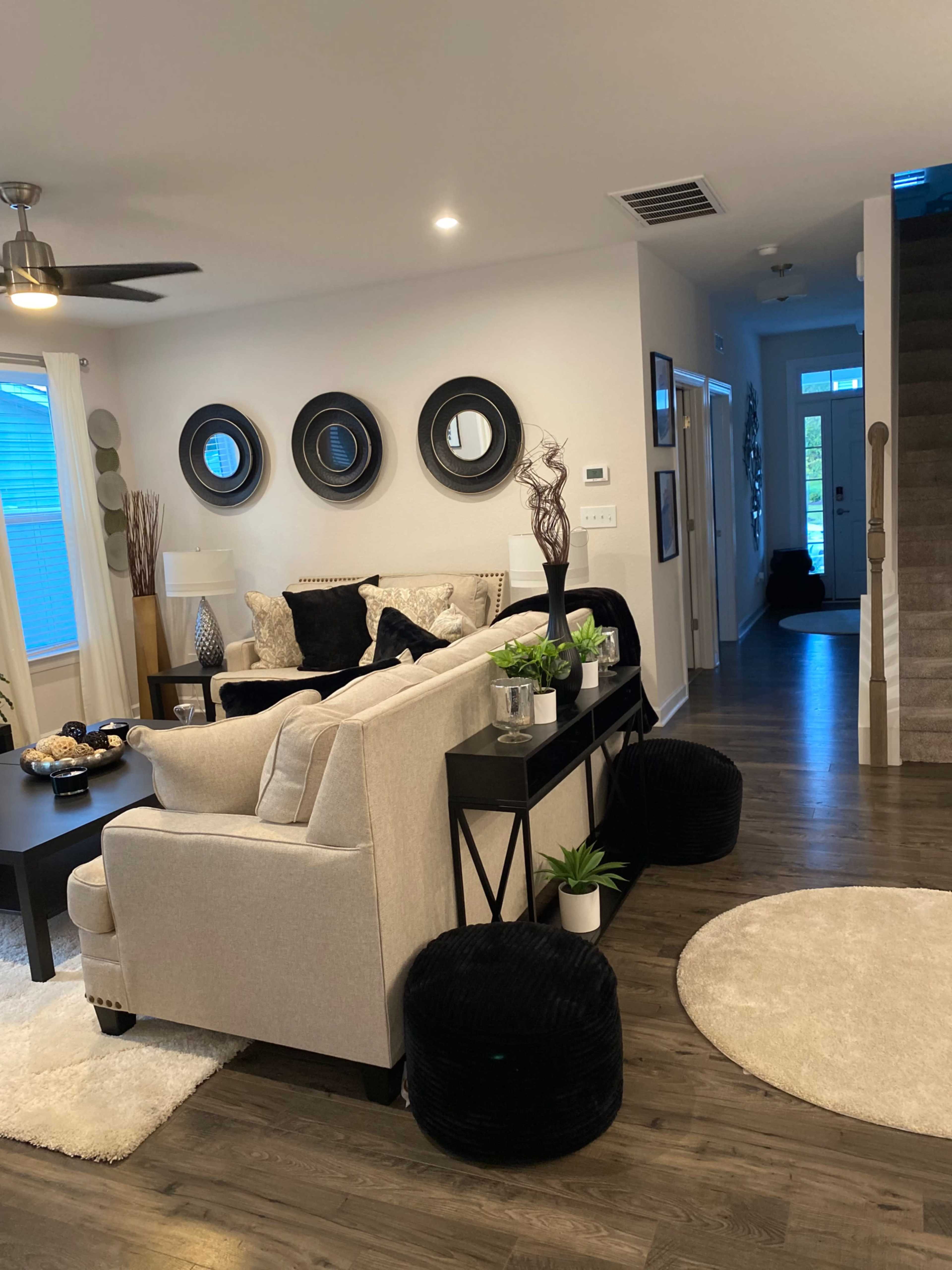 A modern living room with a light-colored sofa, decorative mirrors on the wall, and a console table displaying plants and decor, leading to a staircase and hallway.